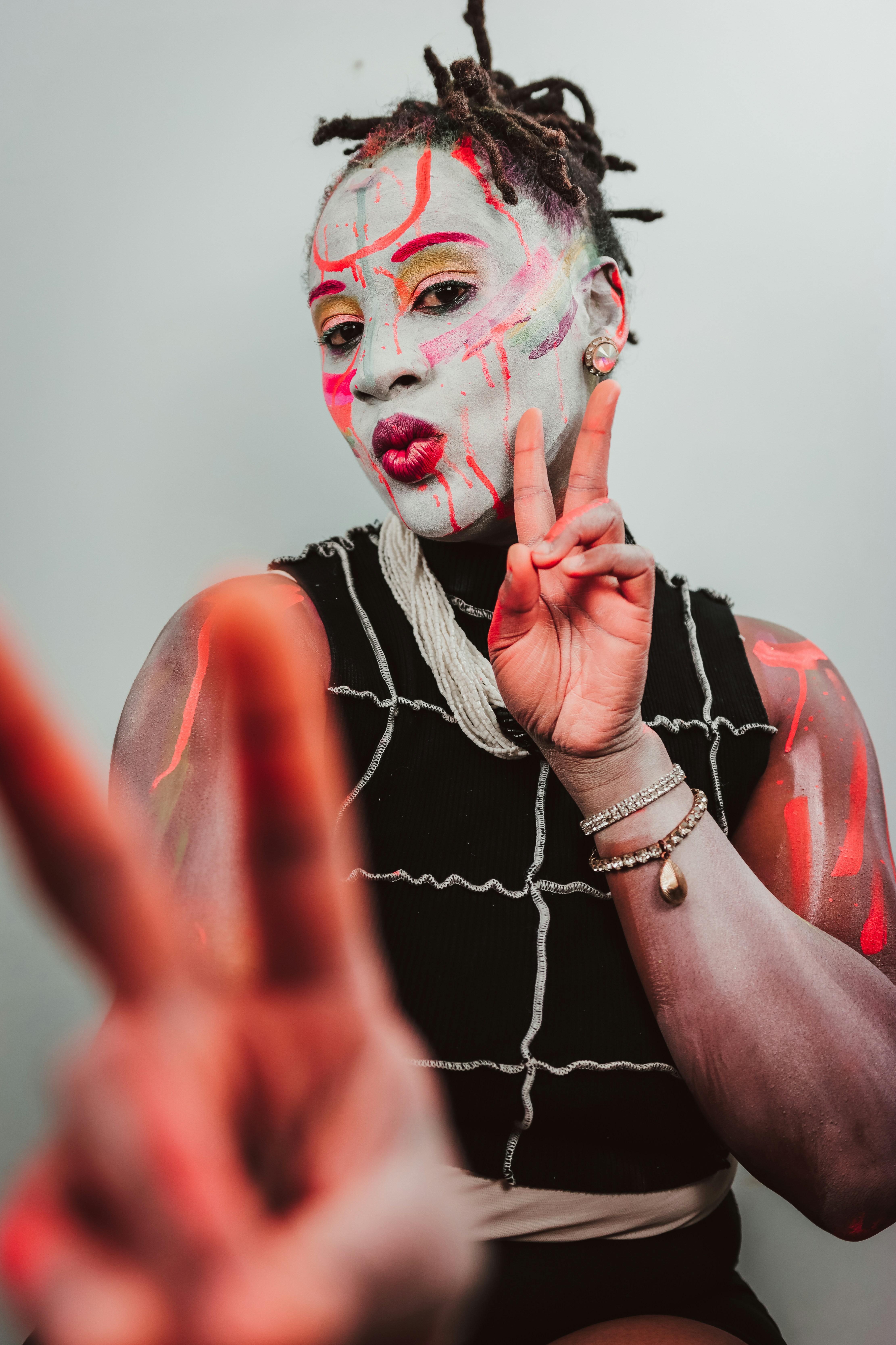 A person with white face paint and neon markings making a peace sign gesture