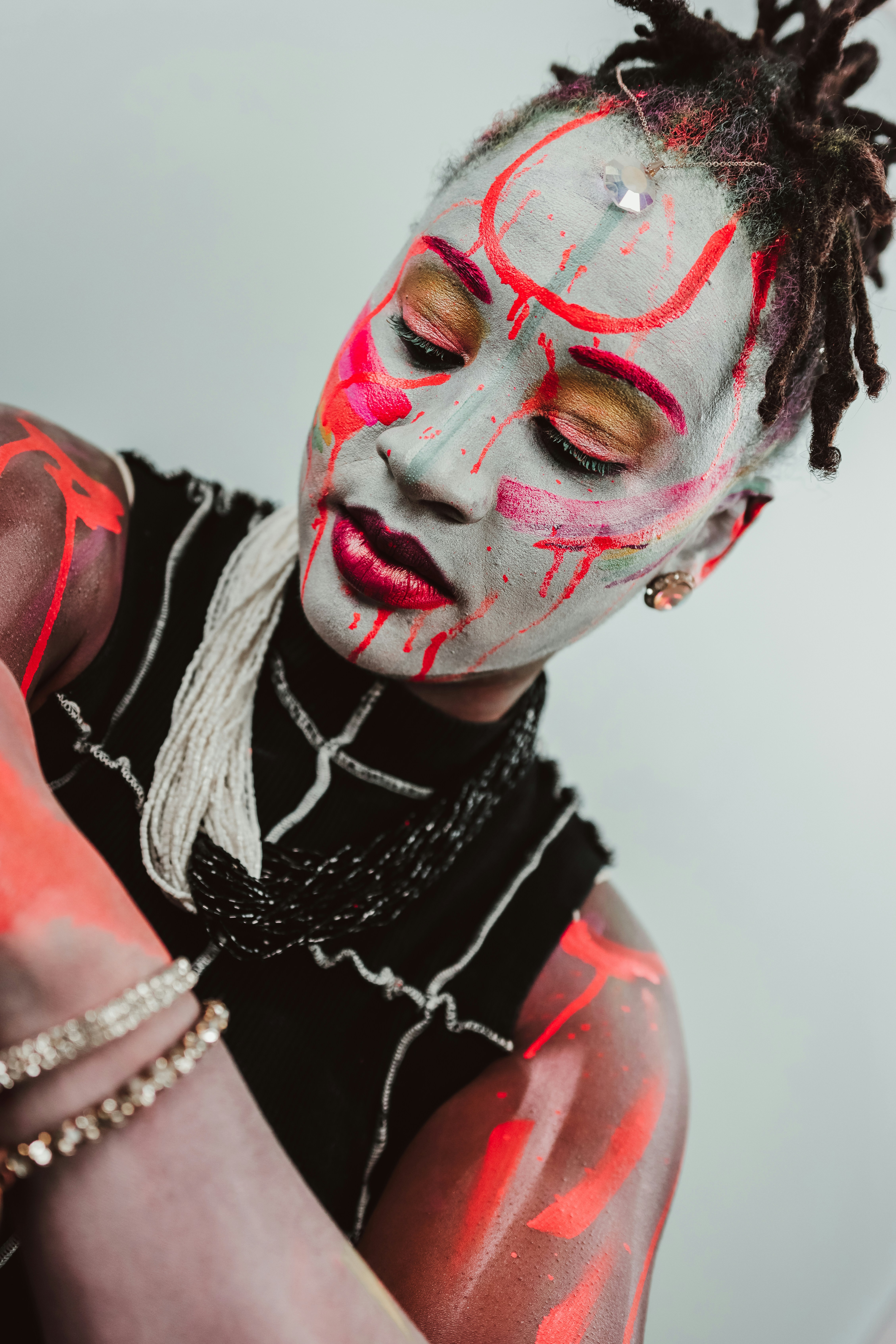 A person with grey face paint and red abstract markings wearing a black top and beaded necklaces