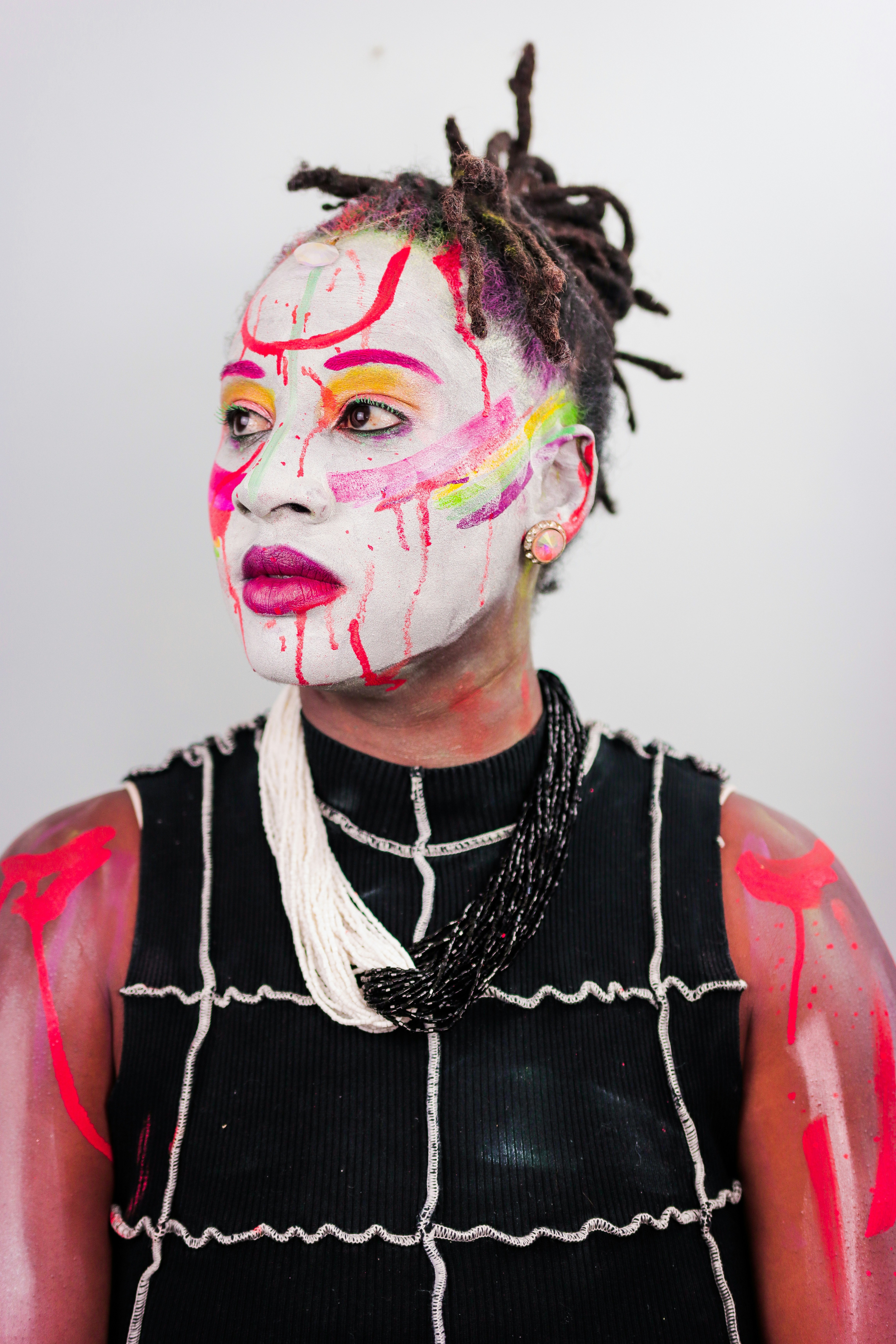 A person with white face paint and colorful abstract markings wearing a black top and beaded necklace