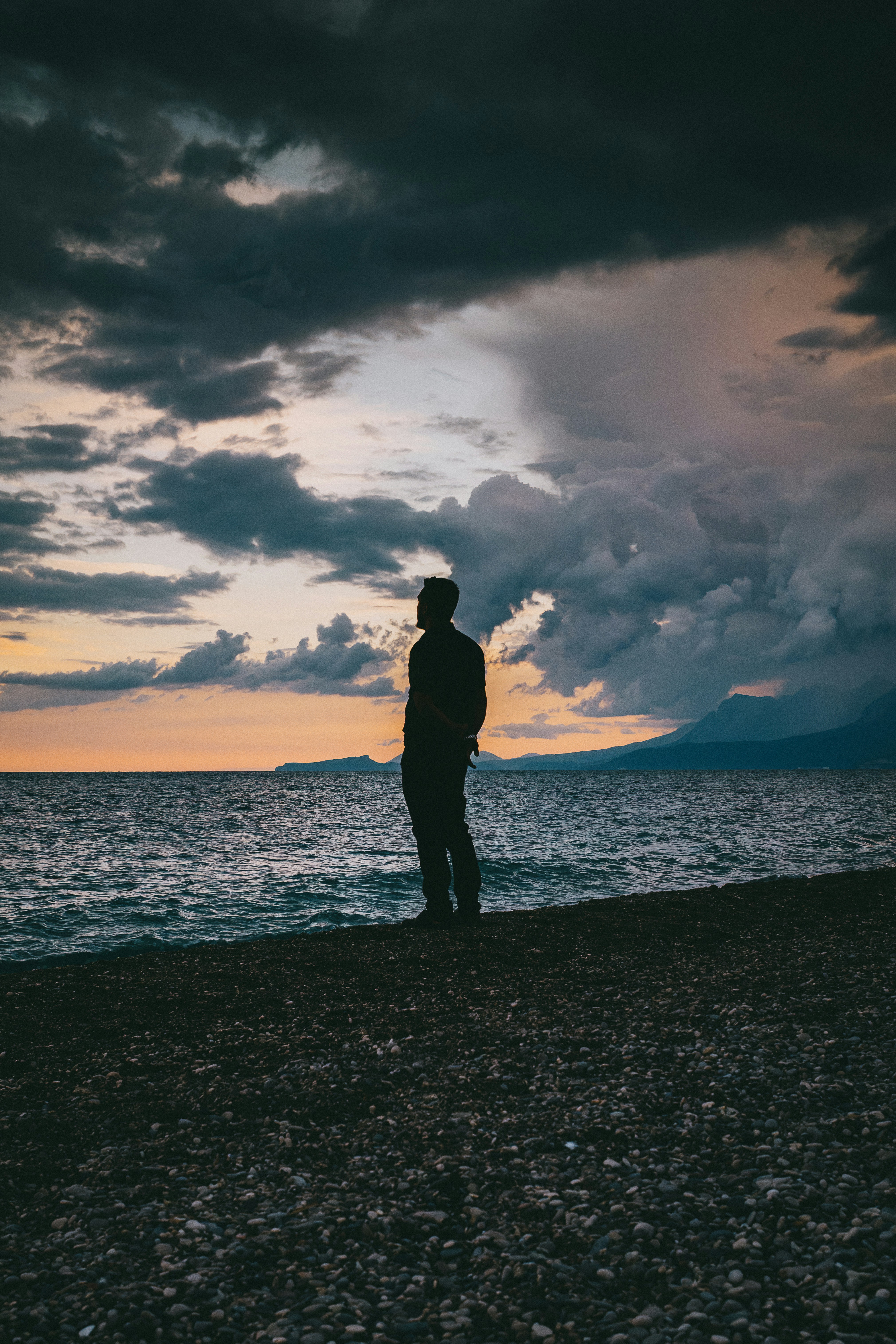 A person in silhouette on a pebble beach against a dramatic cloudy sky at low sun