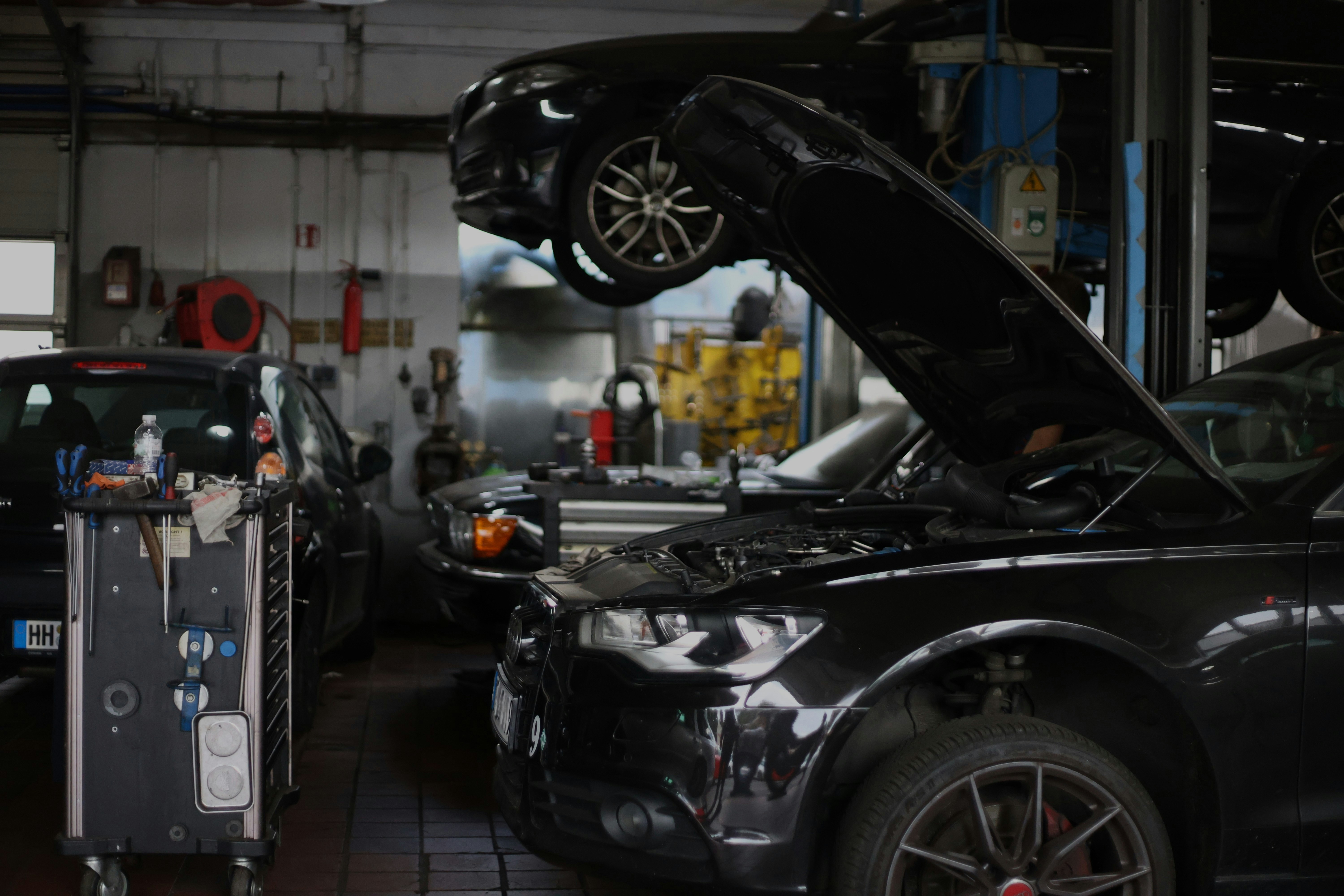Inside a repair shop, a dark hatchback with its hood propped open in the foreground, another car raised on a lift above it and a rolling tool cart at the left