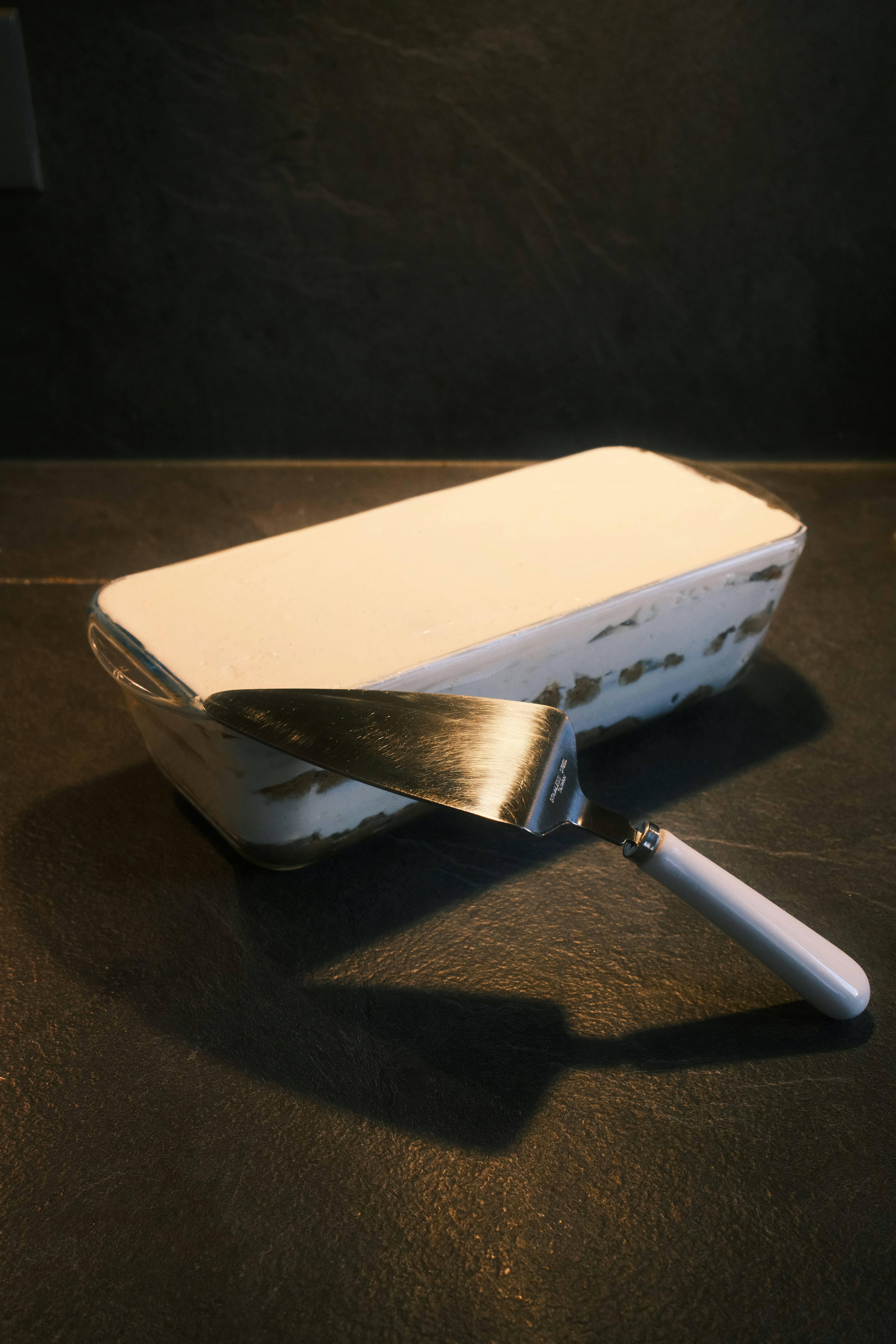 A glass dish of tiramisu with a metal cake server on a dark textured surface