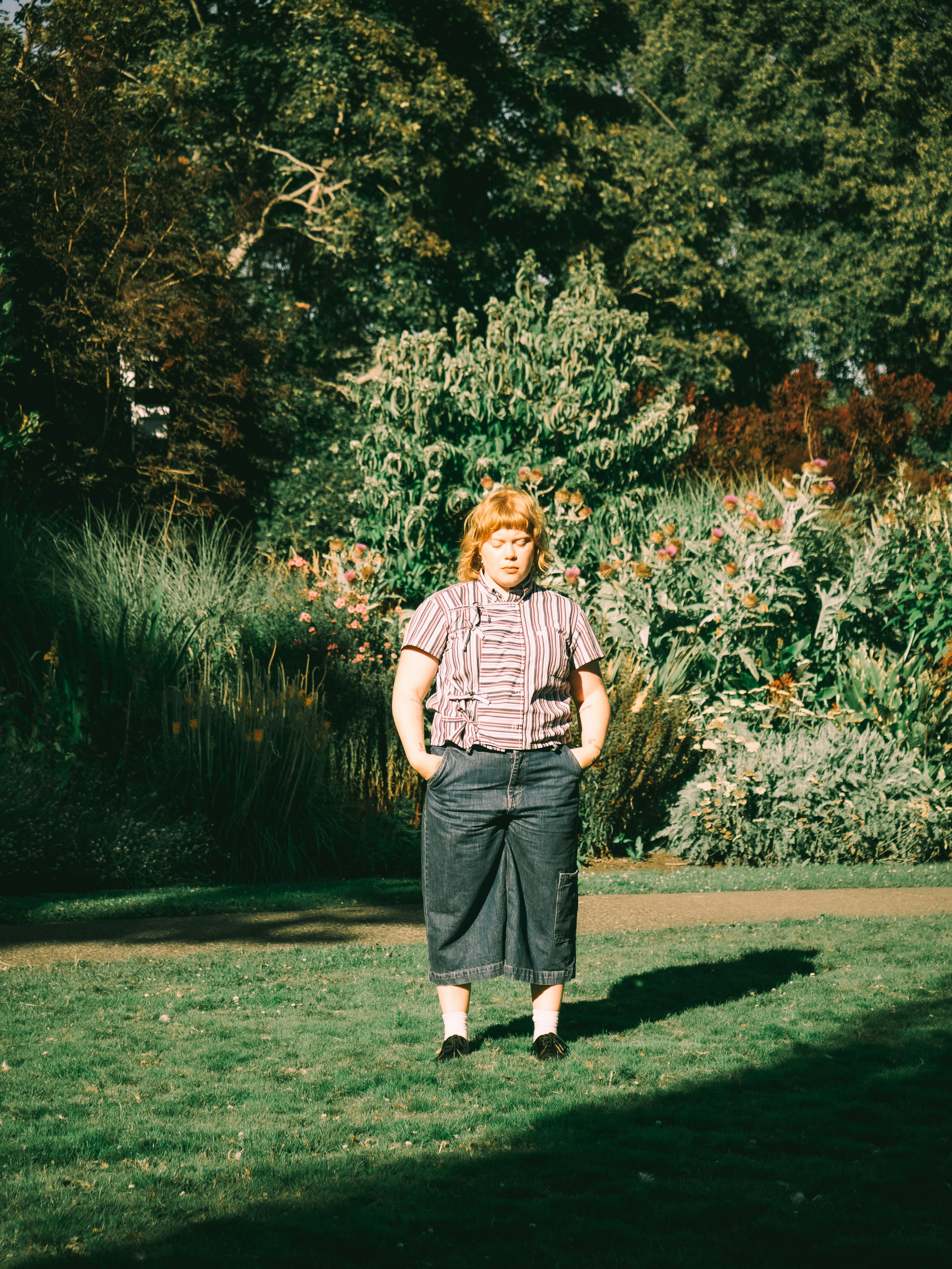 An adult in a striped shirt and denim culottes standing in a sunlit garden