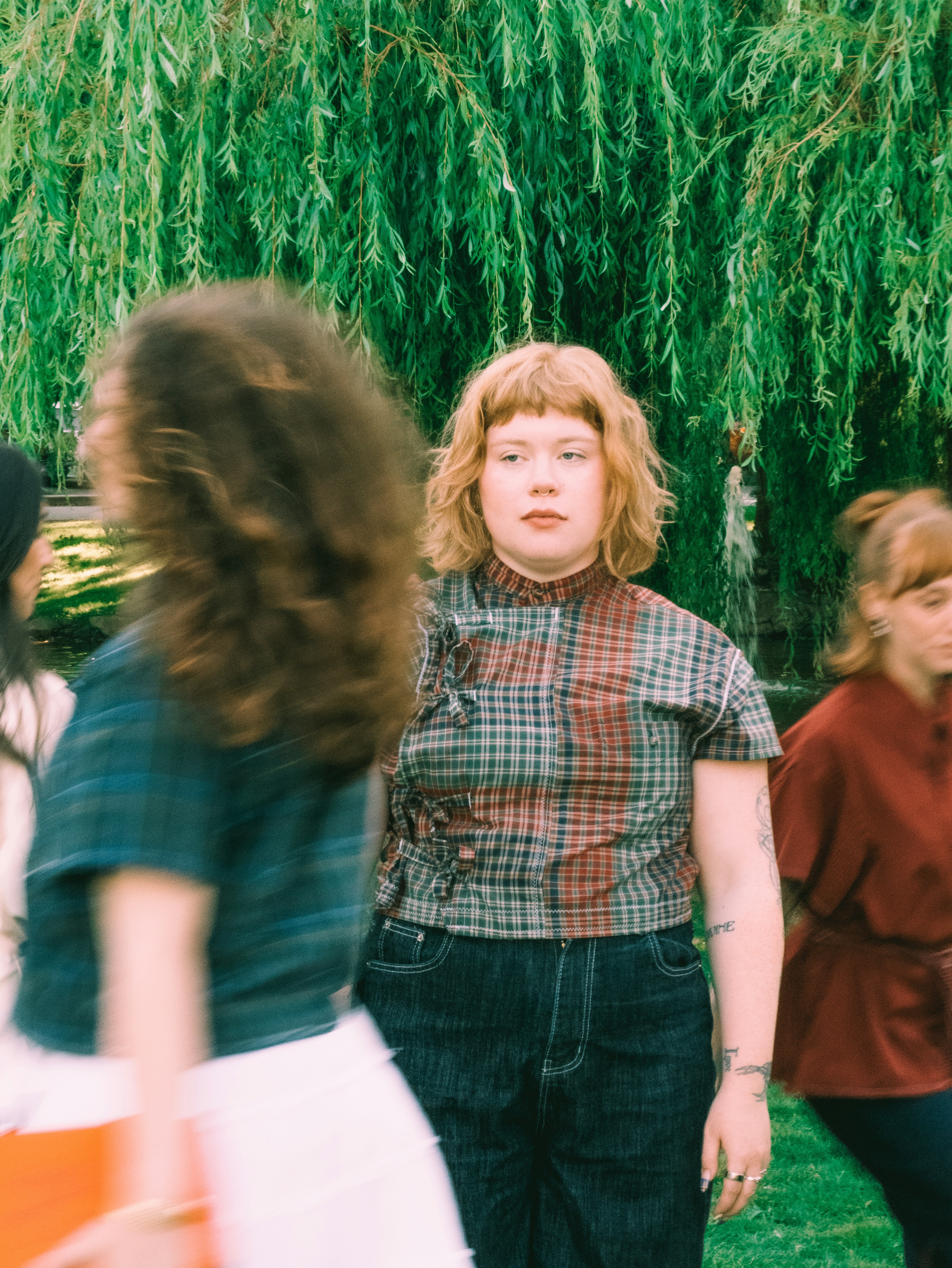 A woman with short ginger hair in a plaid top standing before a weeping willow