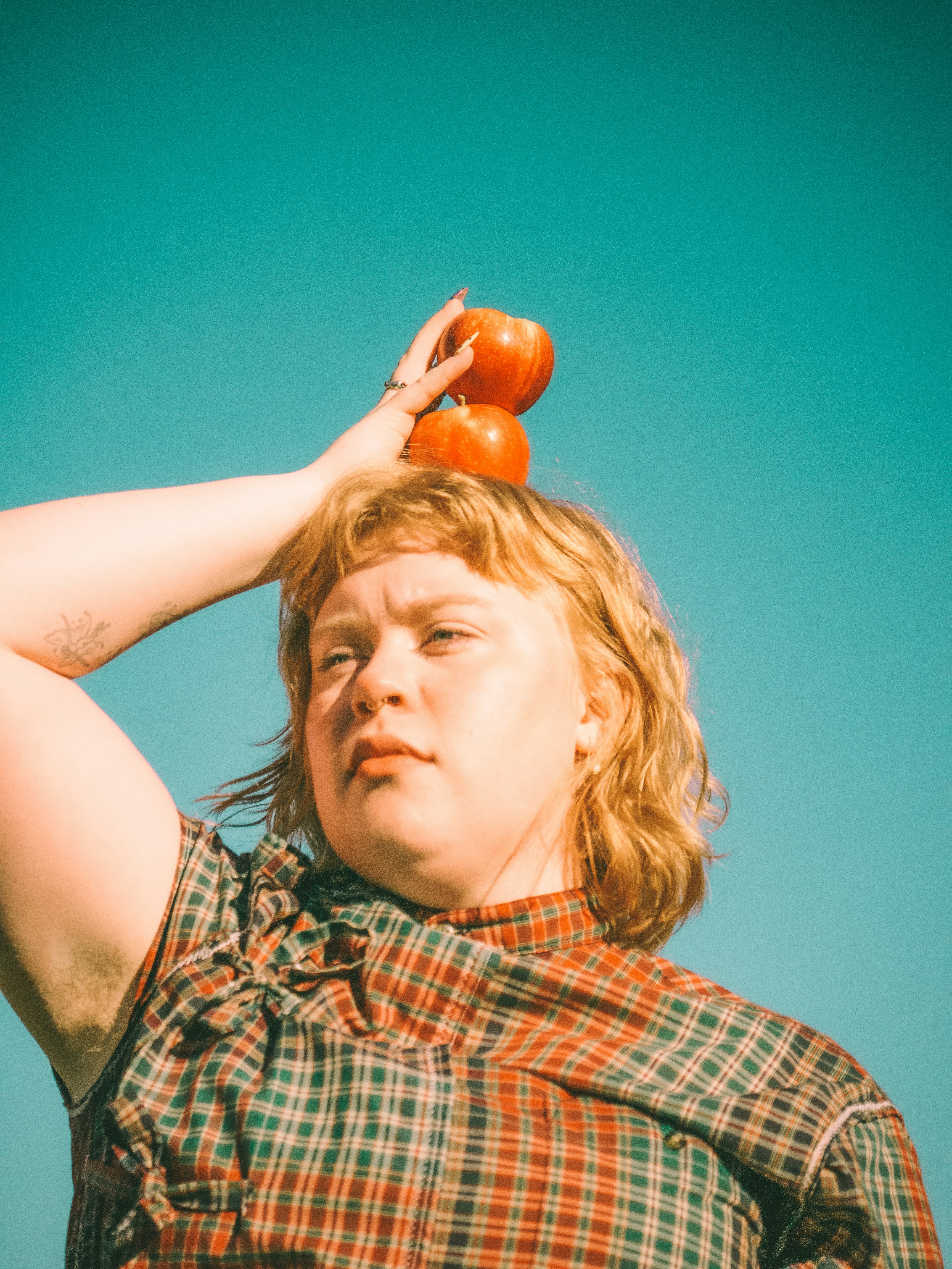 A person in a plaid shirt balancing two red apples on their head against a bright teal sky
