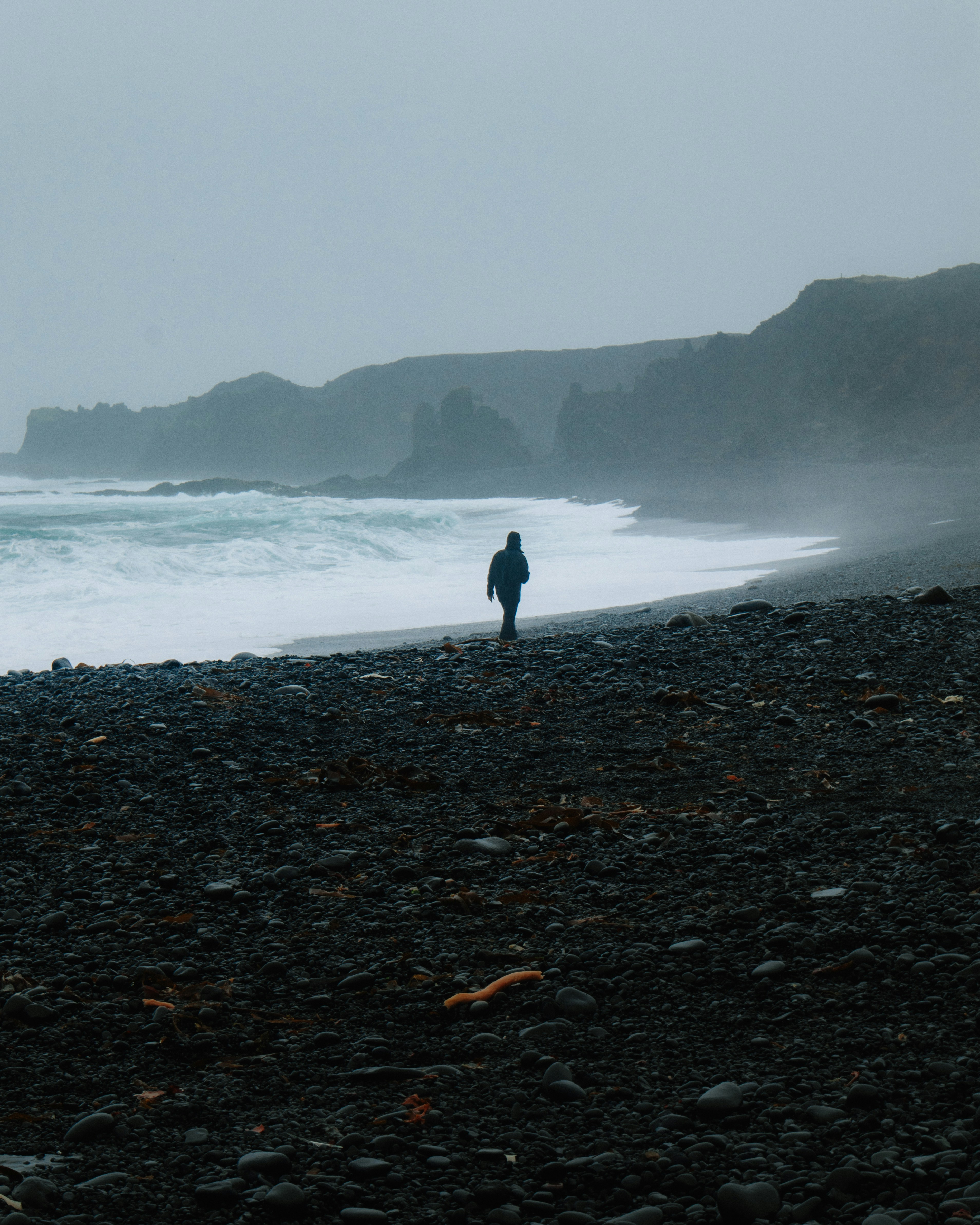 A person walking along a misty black pebble beach beside a crashing ocean surf