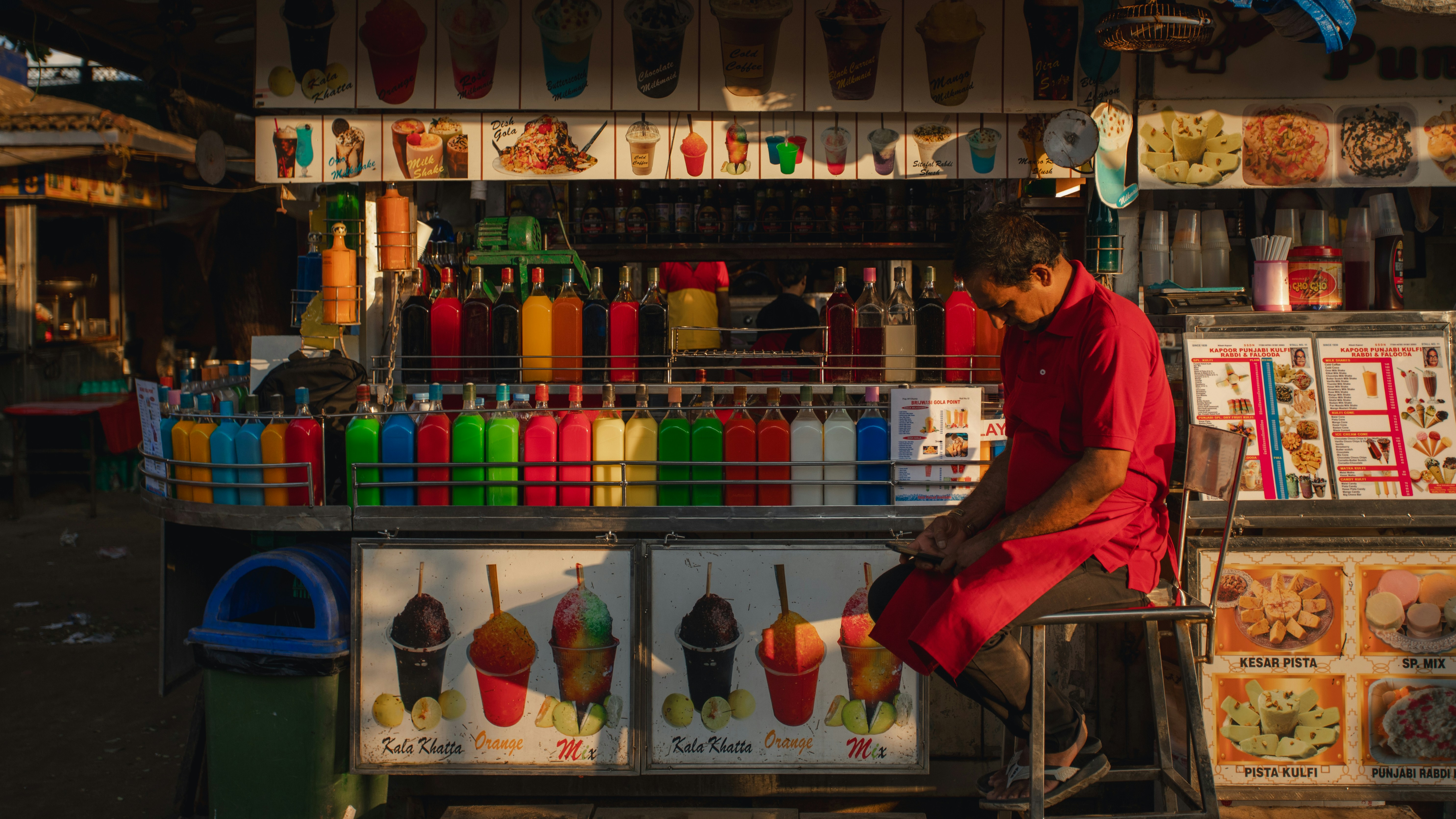 A man in a red shirt sitting at a colorful street food stall with many bottles