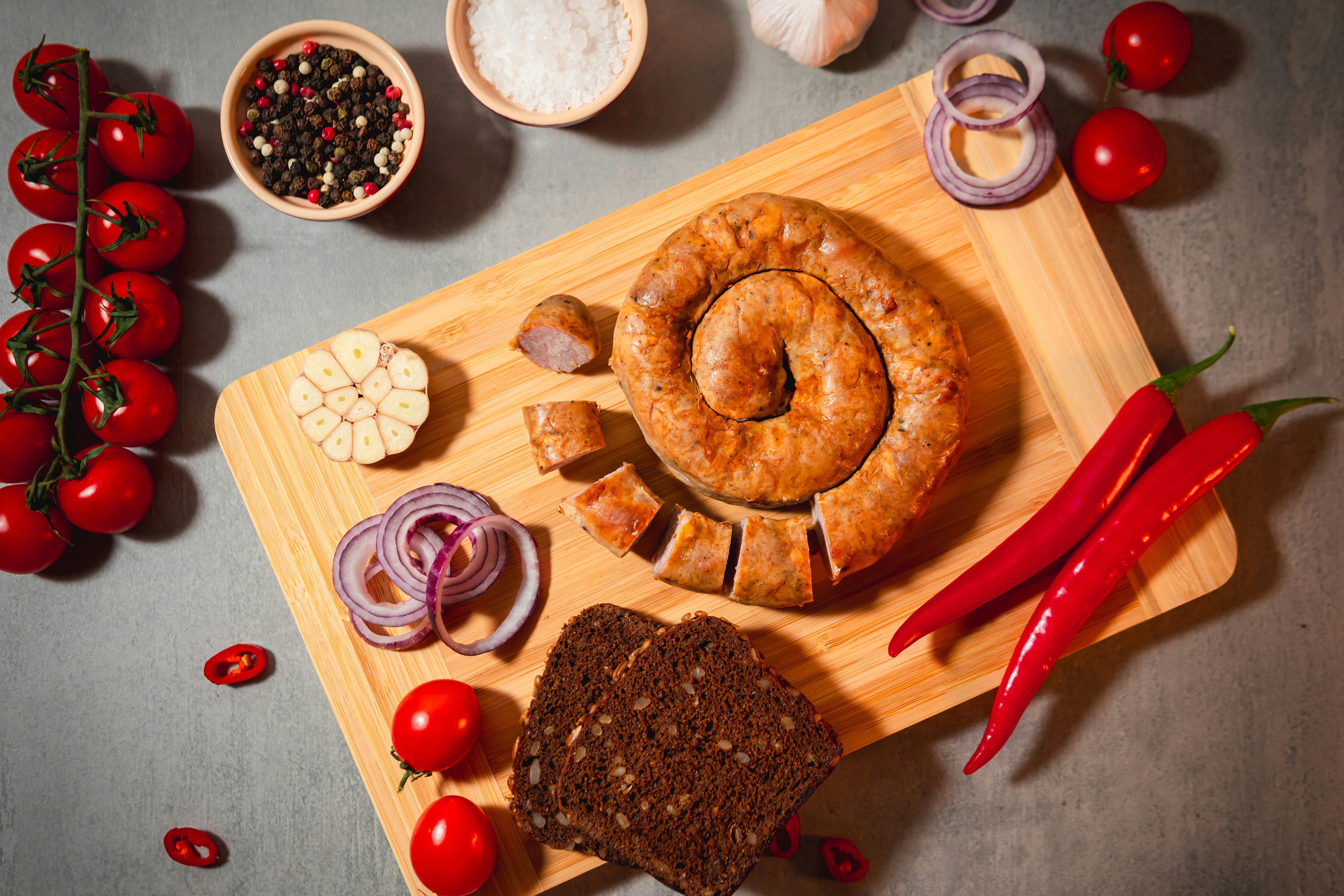 A coil of cooked sausage on a wooden cutting board with sliced bread, cherry tomatoes, and red onions