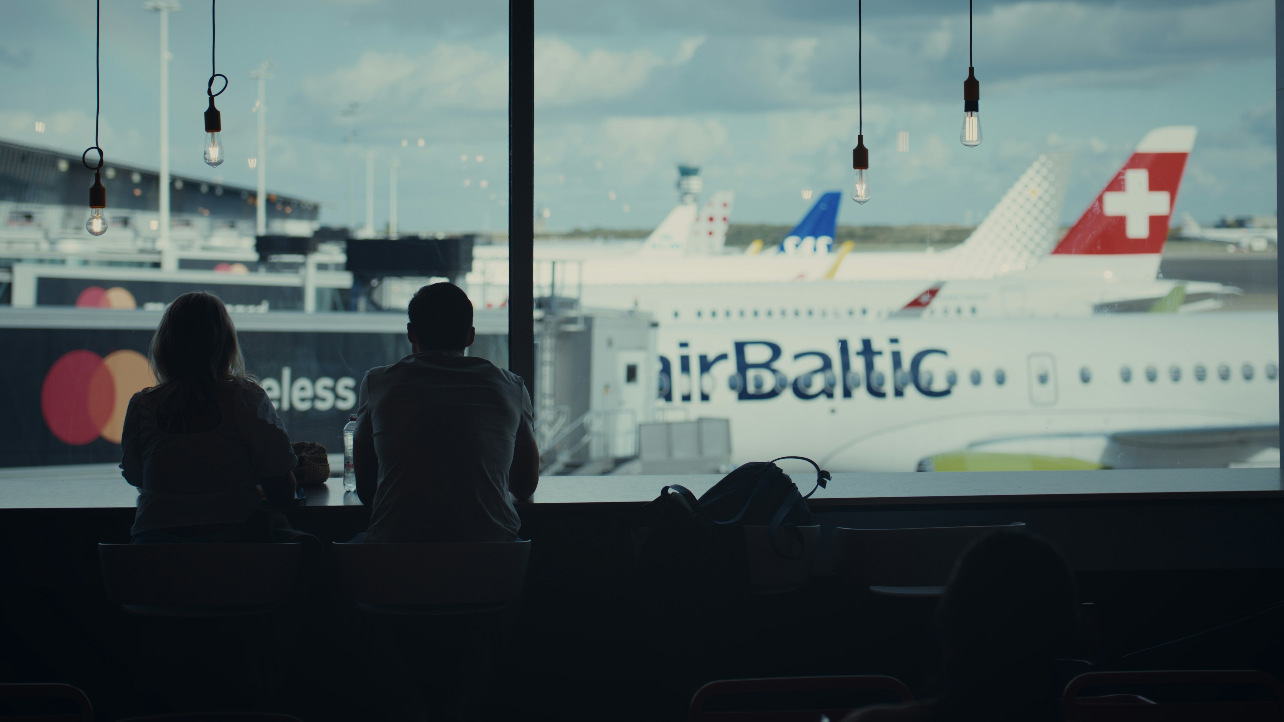 Two people sitting at an airport terminal window watching airplanes on the tarmac