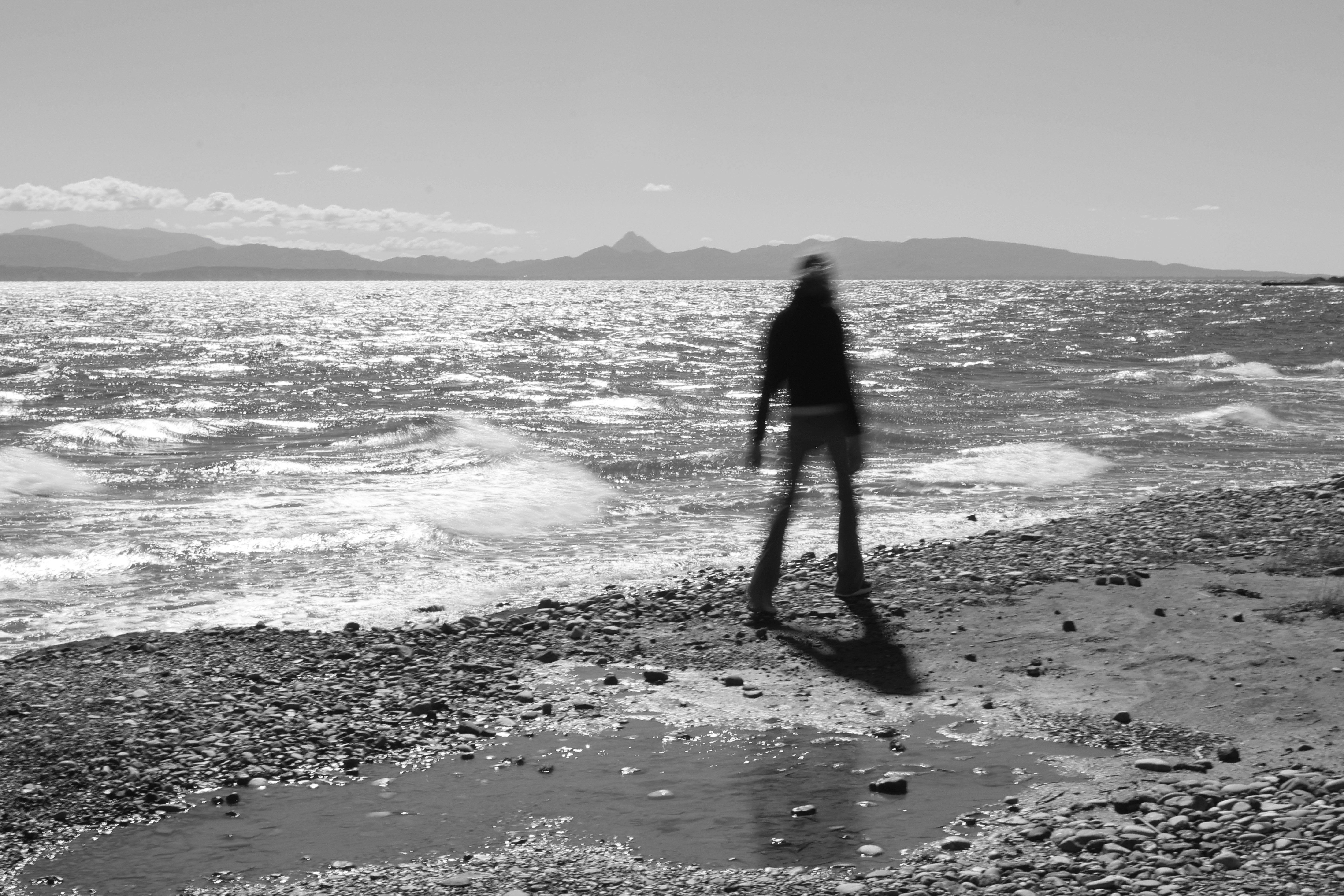 A blurred person walking along a pebble beach by the sea in black and white