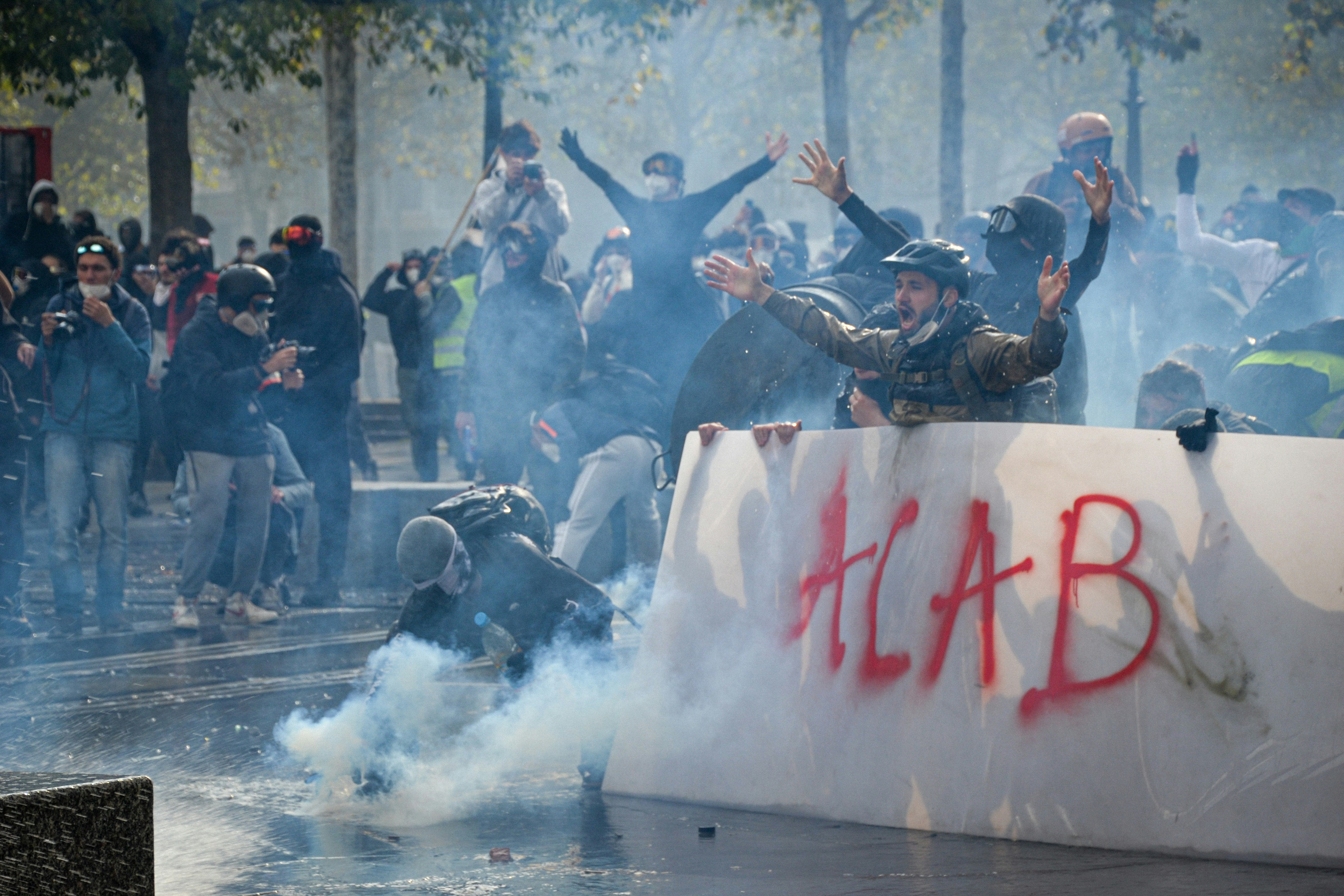 Protesters behind a white barrier with red graffiti in a cloud of tear gas