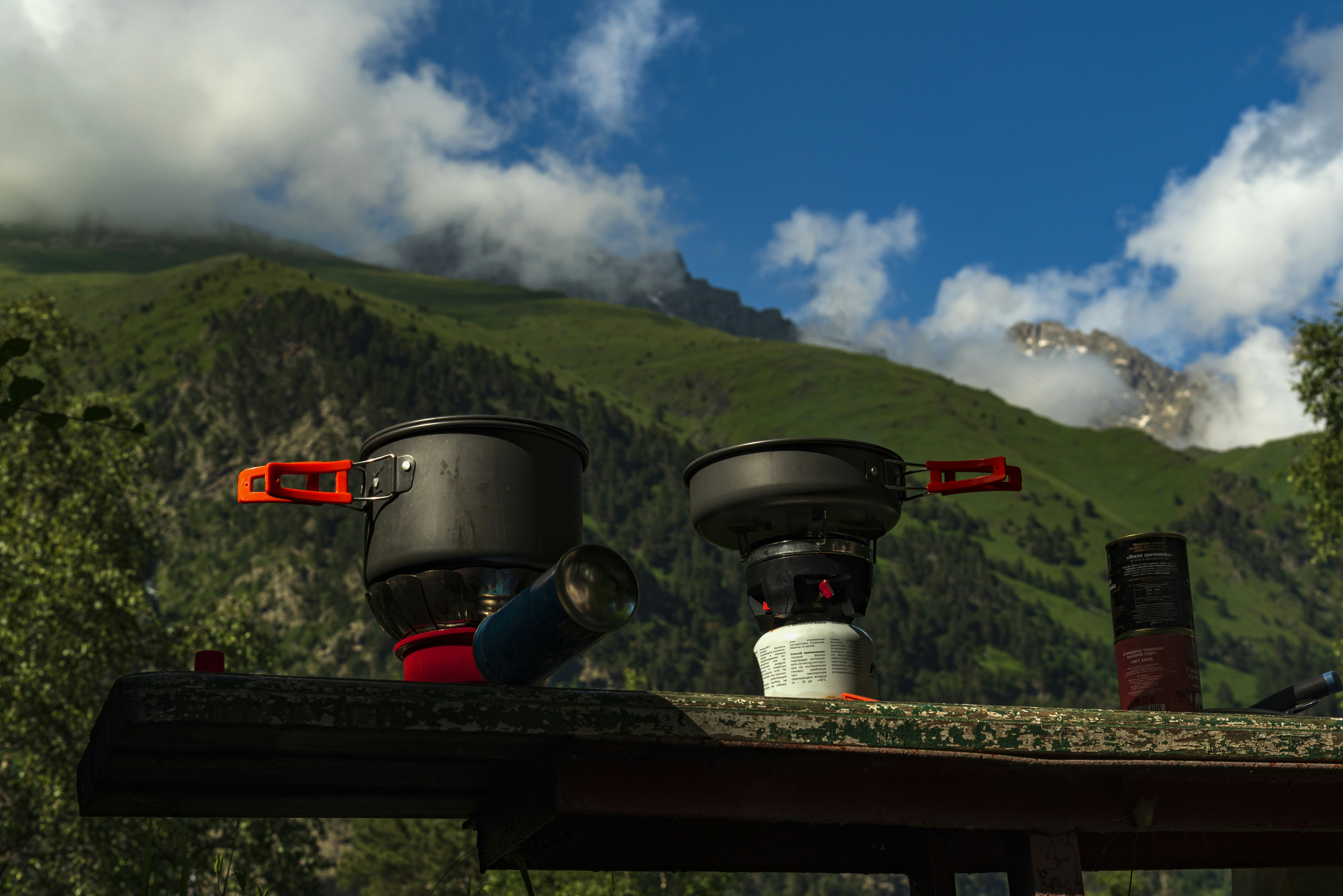 Two camping stoves with cook pots on a wooden table in front of green mountains
