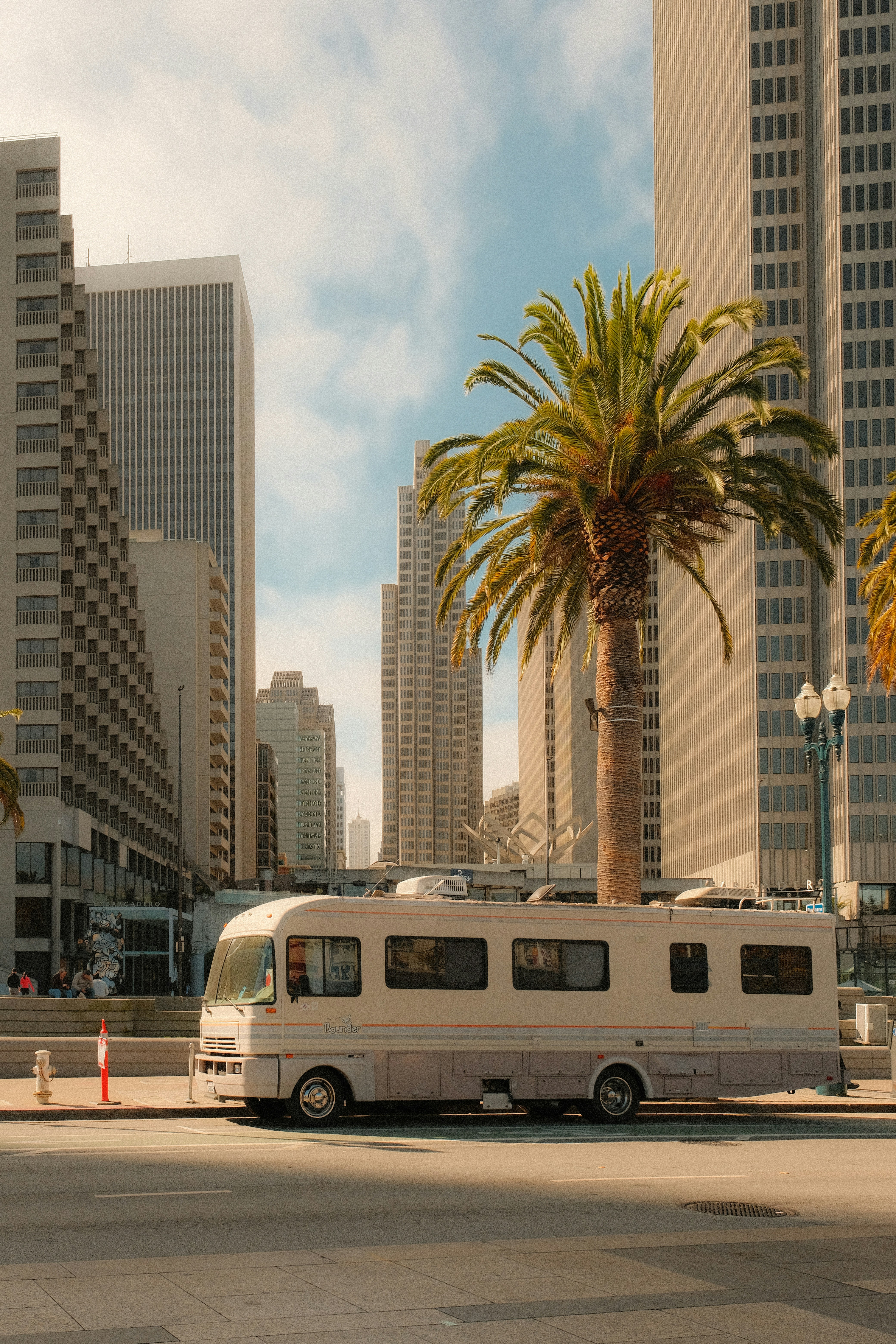 A recreational vehicle parked on a city street beside a tall palm tree and skyscrapers