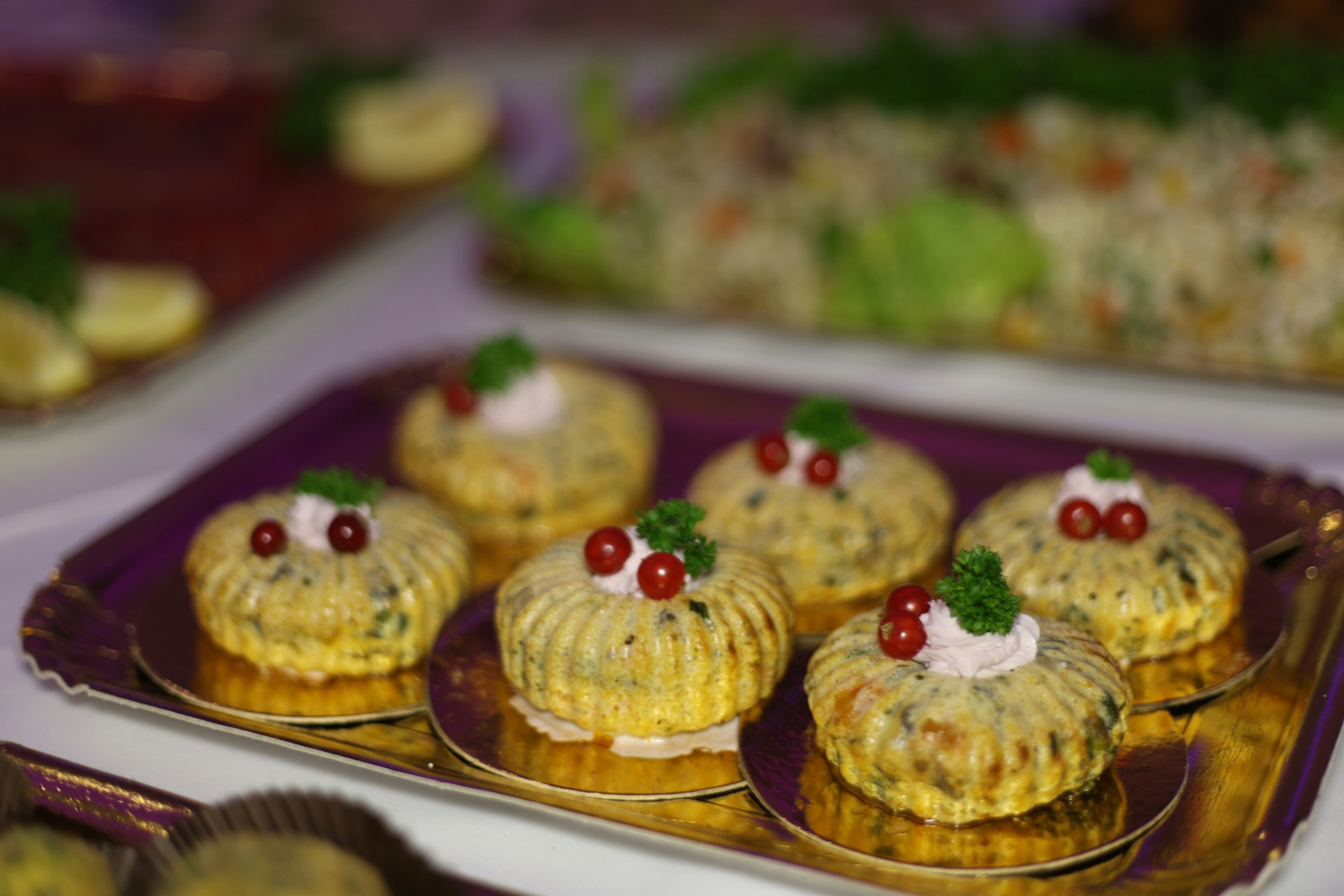 A beautifully styled food photograph featuring delicate, bite-sized appetizers arranged on an elegant purple serving tray. Each piece is carefully decorated with a creamy white topping and vibrant red garnishes, creating an eye-catching presentation. Soft background blur and warm lighting highlight the intricate textures, colors, and details of these handcrafted culinary creations. Photography by Xalo Mustafa.