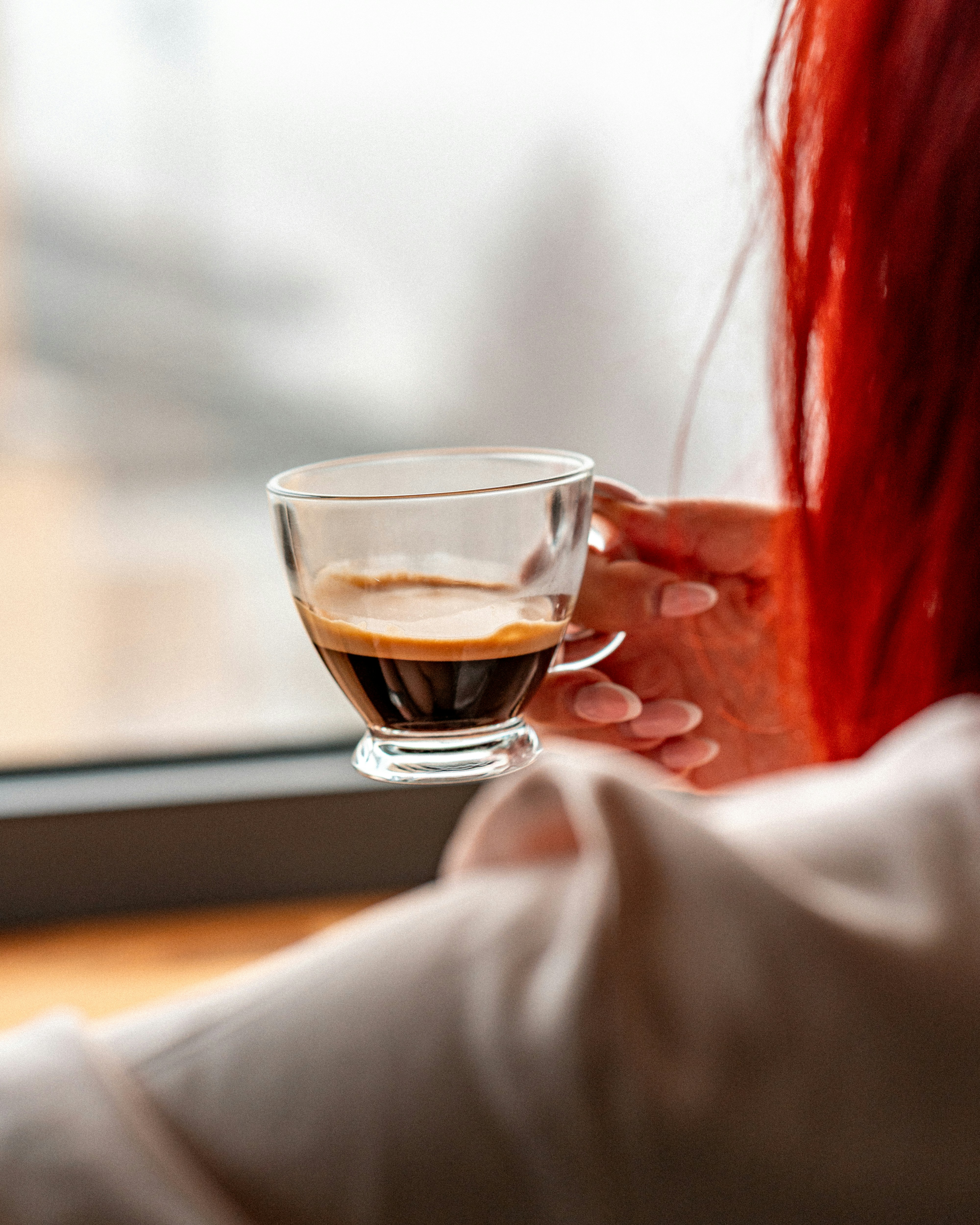A person with bright red hair holding a glass cup of espresso by a window