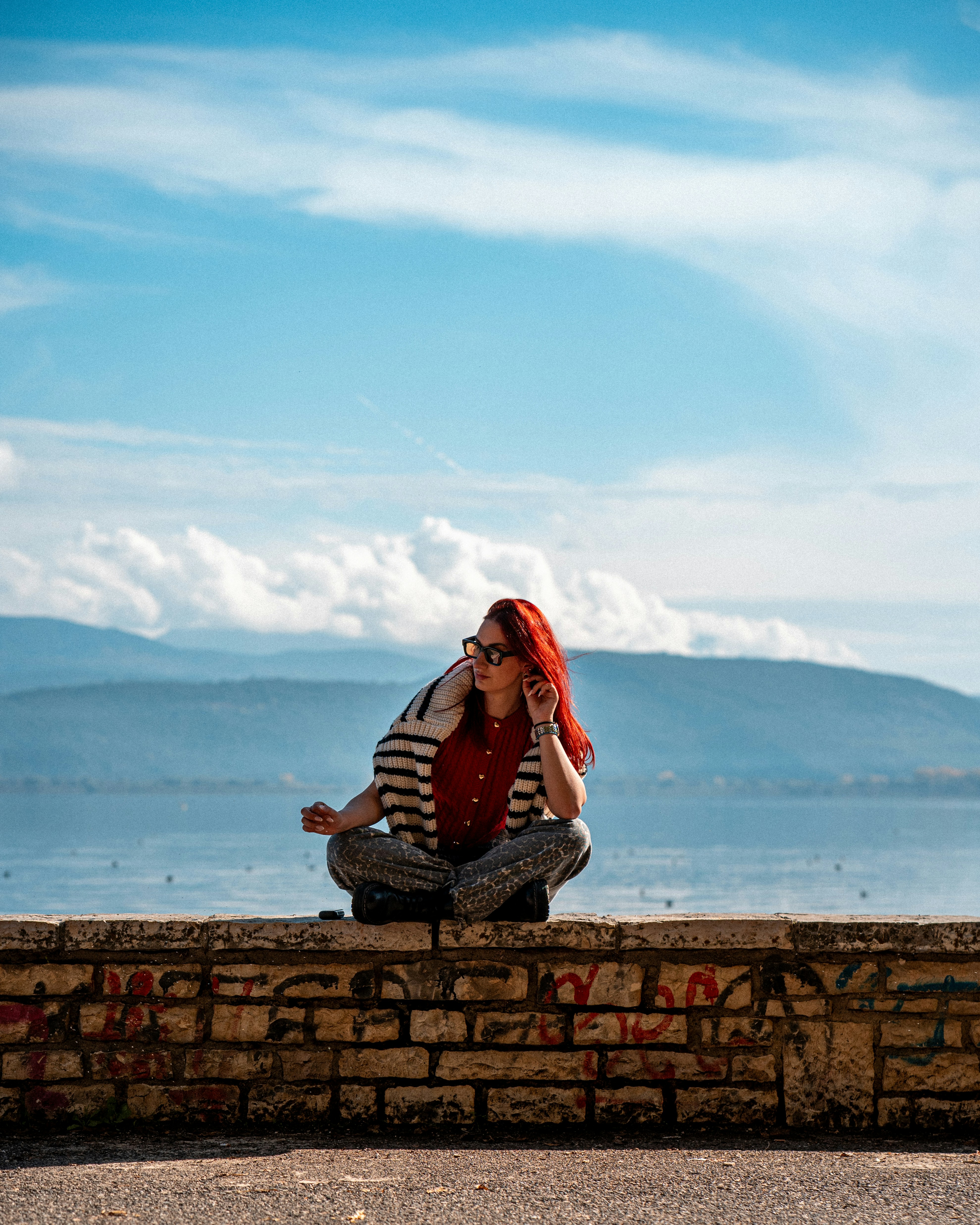A woman with red hair sitting cross-legged on a stone wall overlooking a lake