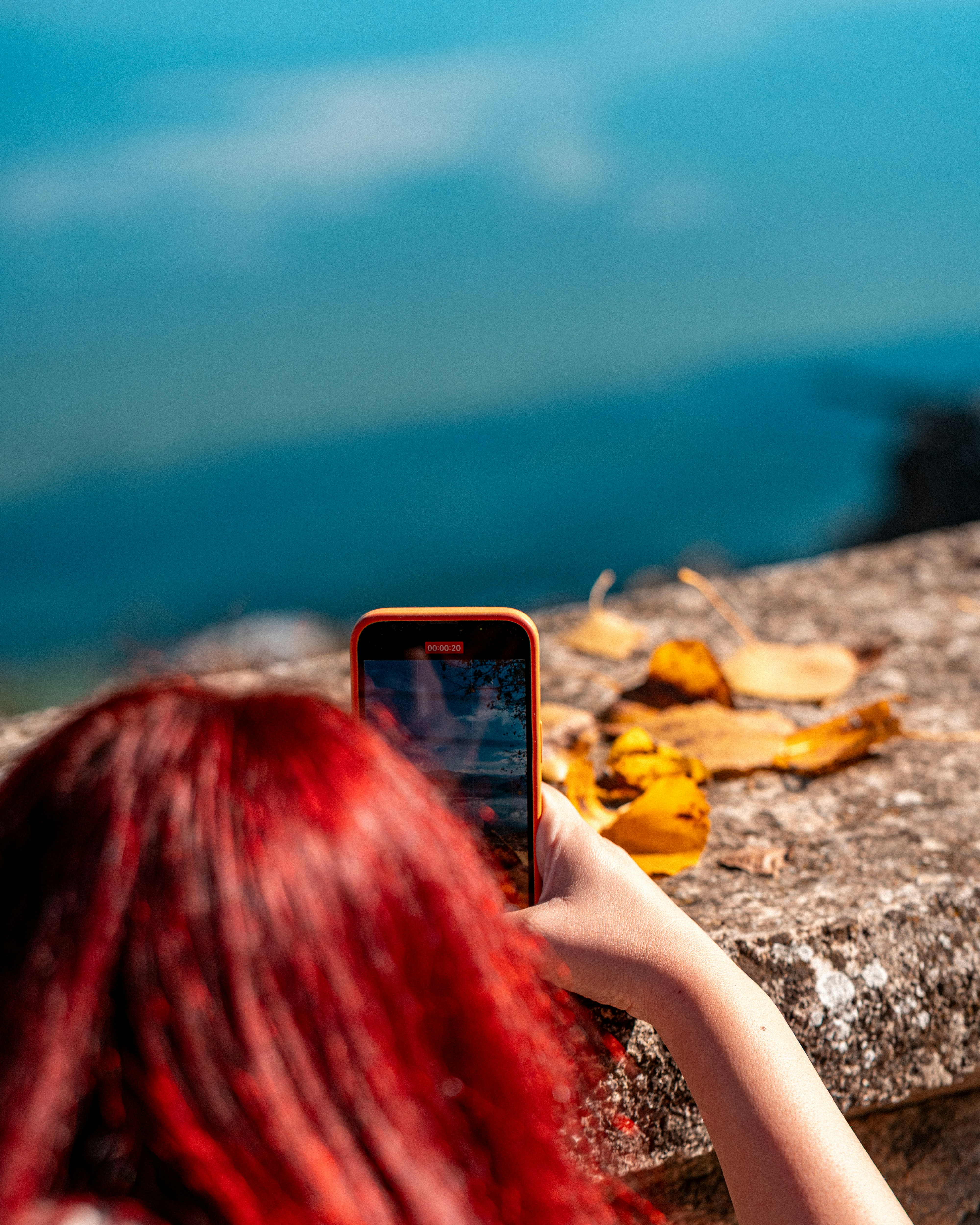 A person with red hair filming a blue landscape with a smartphone on a stone wall