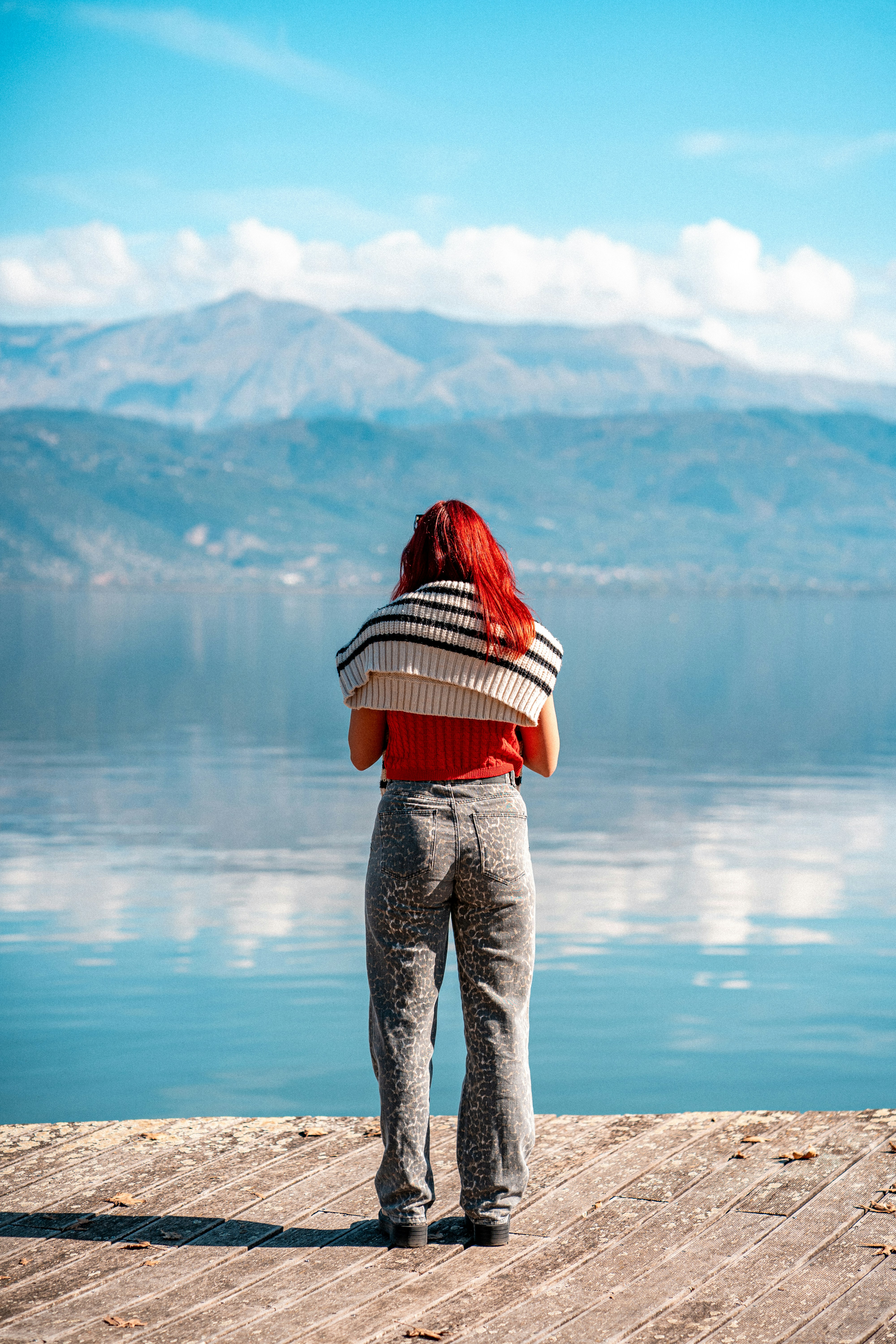 A person with red hair standing on a wooden pier overlooking a calm lake and mountains
