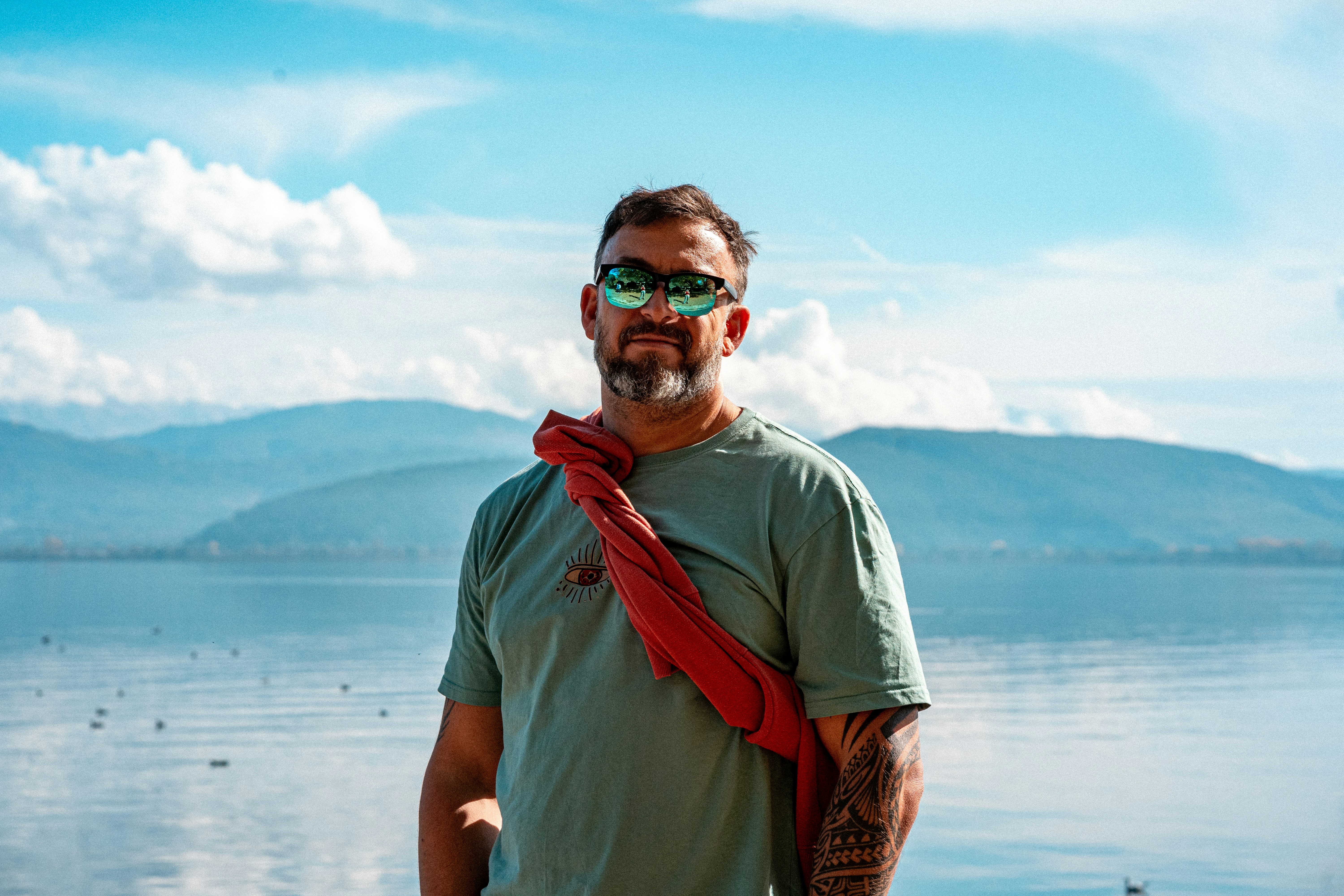 A man with a beard and sunglasses wearing a green t-shirt by a calm lake