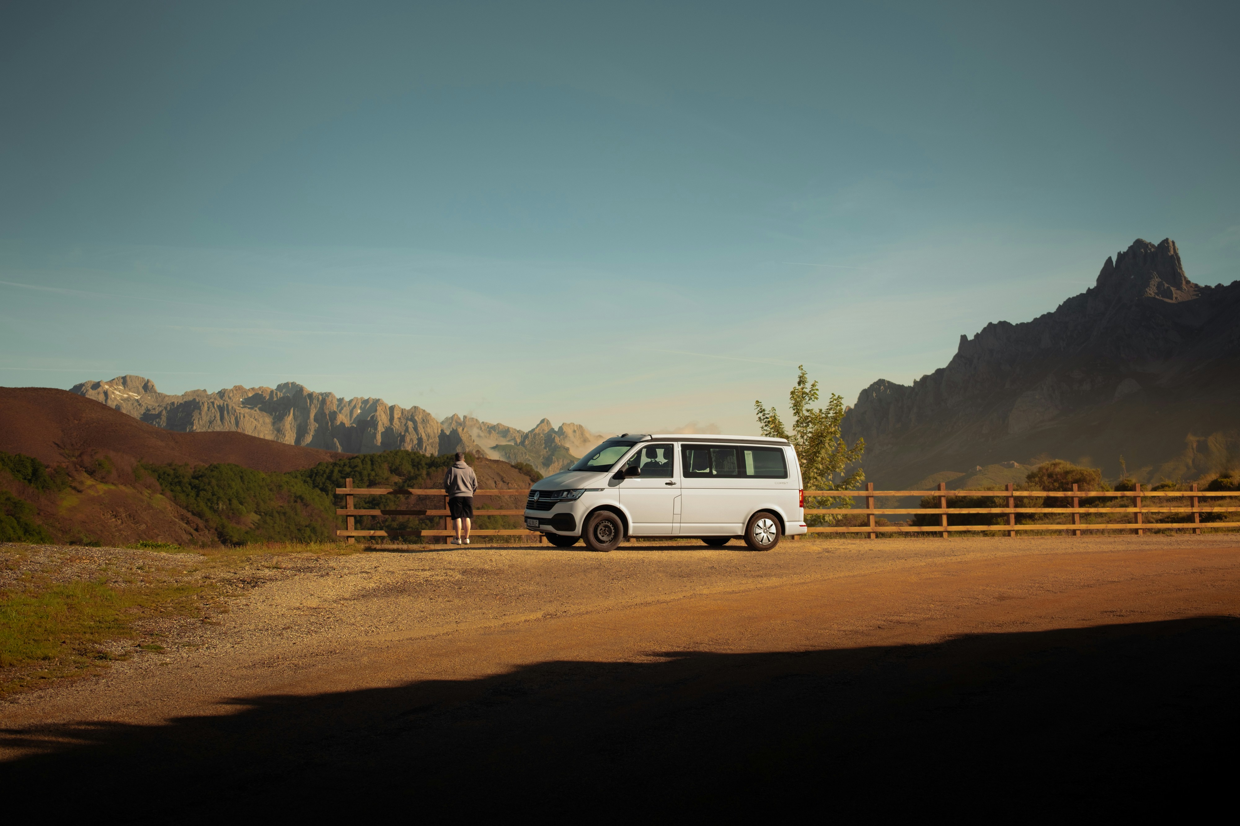 A person standing by a white van parked on a gravel overlook with mountain peaks