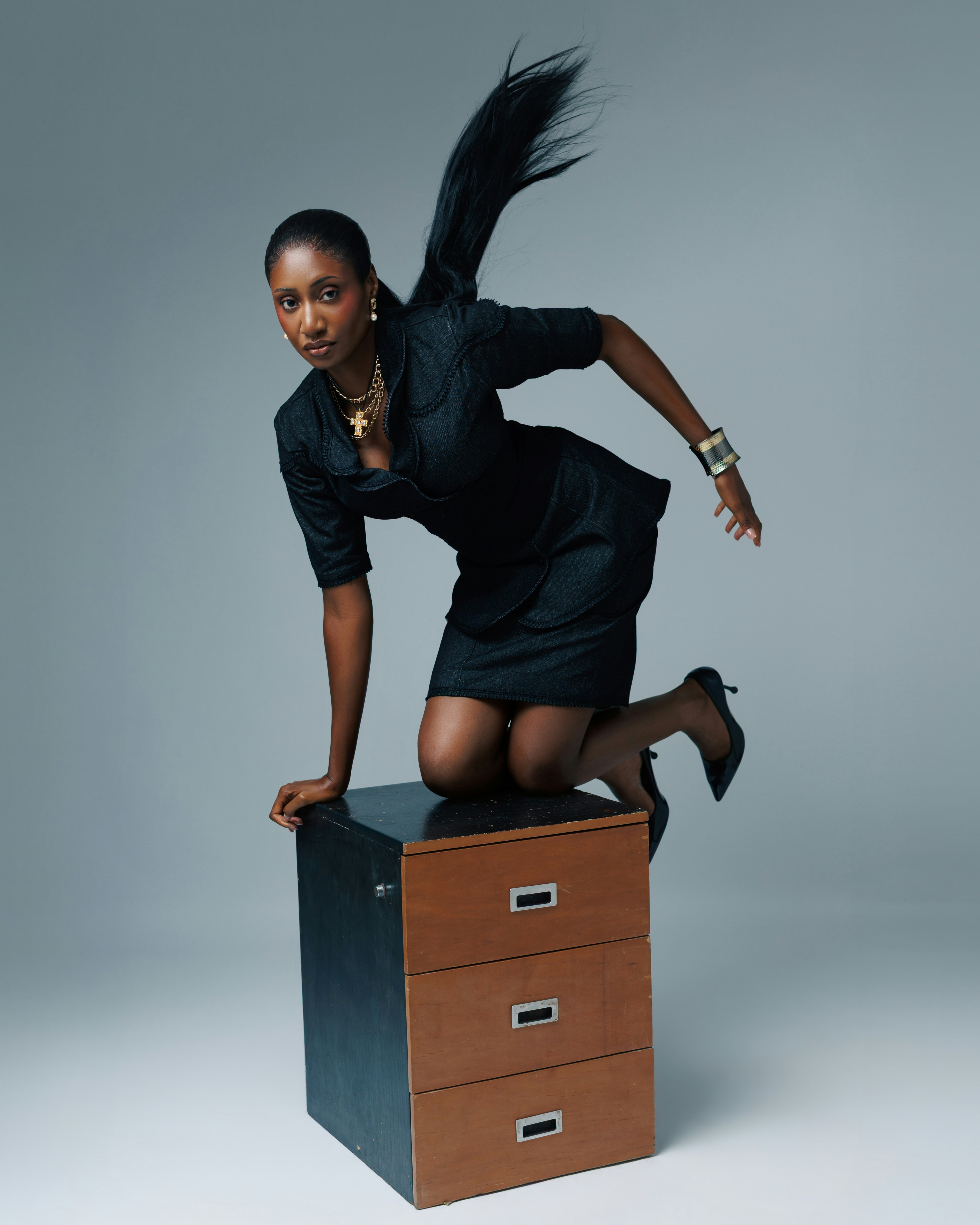 A woman in a dark suit kneeling on a wooden filing cabinet against a grey background