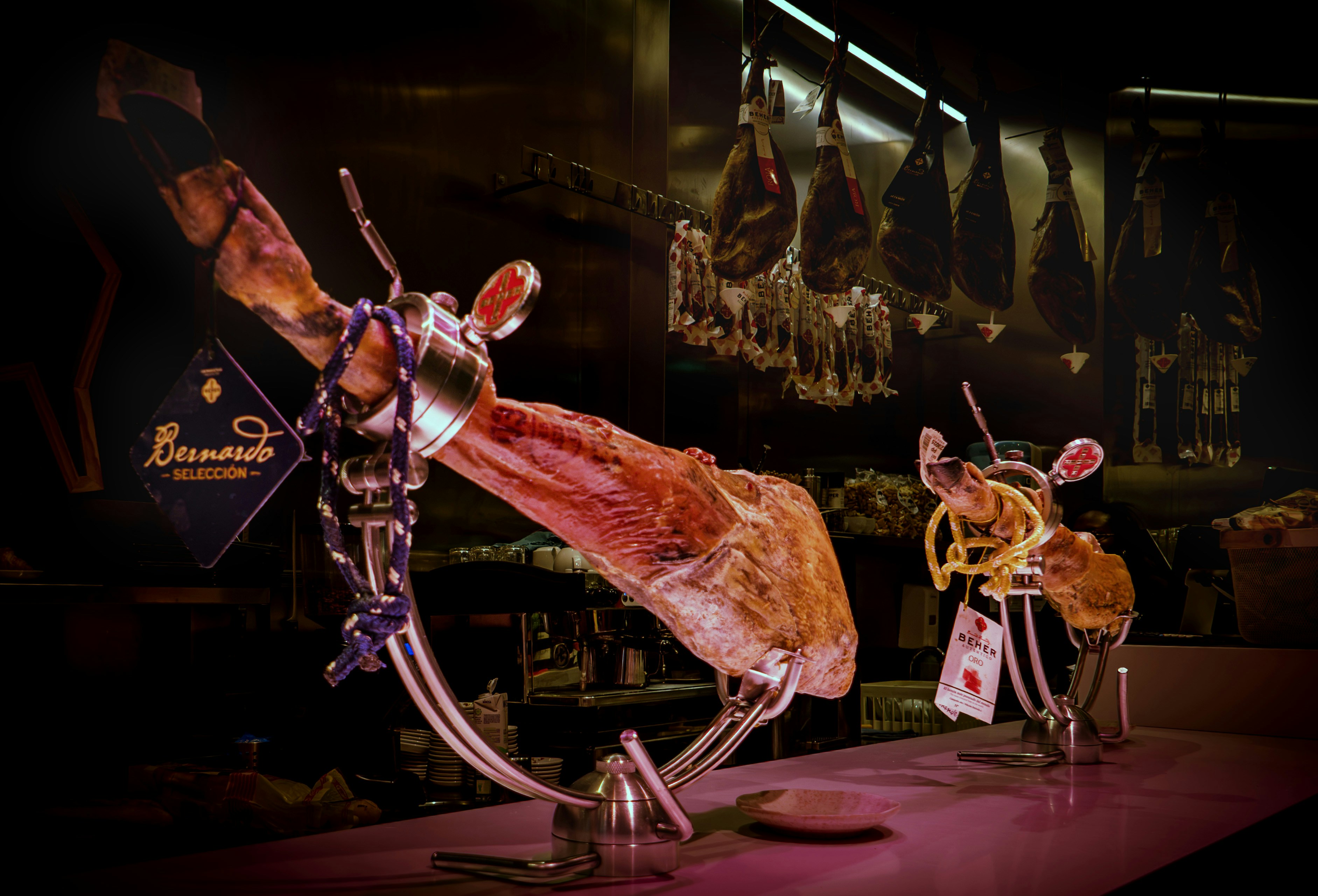 Fotomural Two cured ham legs in metal stands on a counter with hanging hams in the background