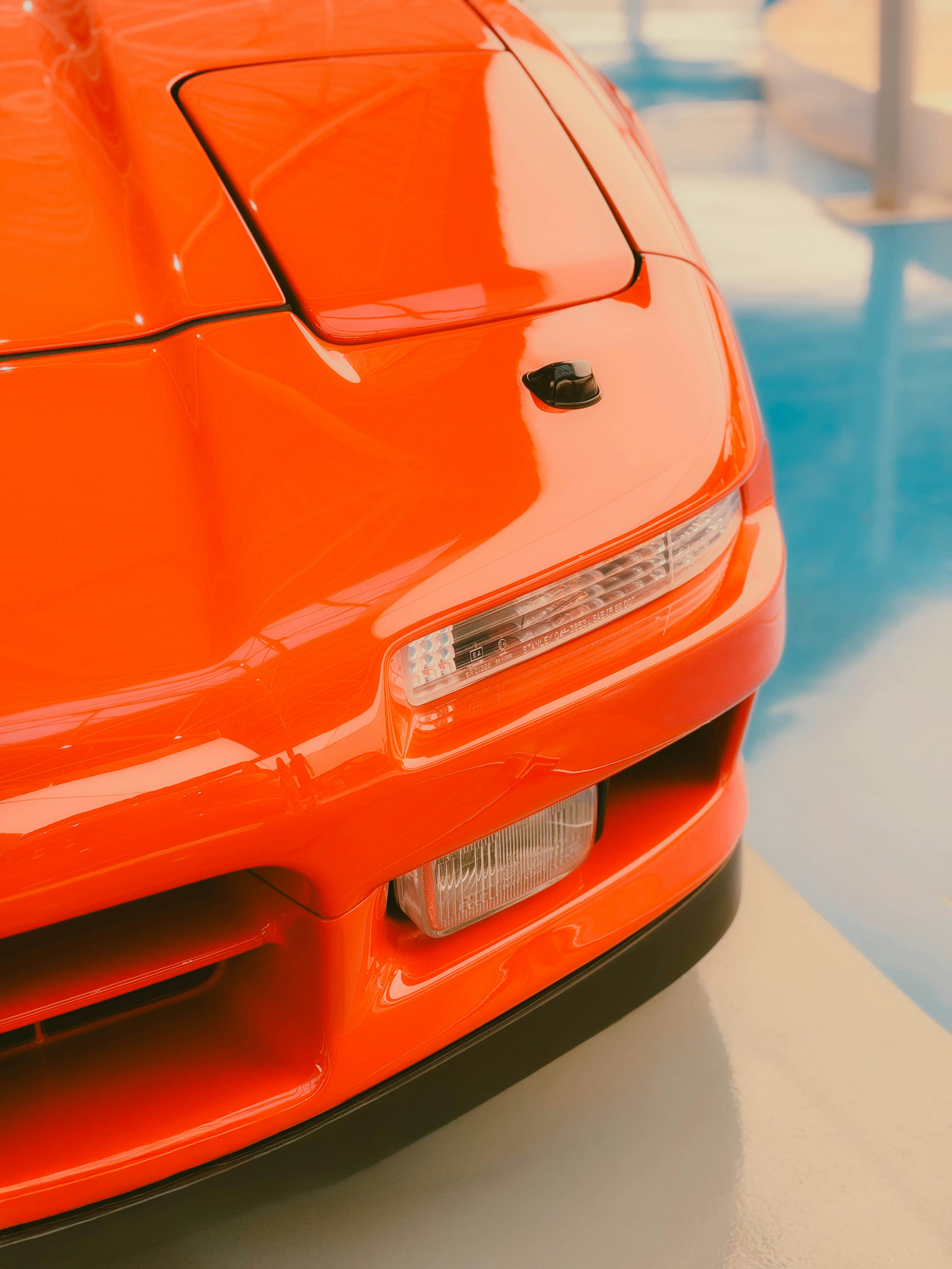 The front corner of a bright orange sports car with pop-up headlights on a blue floor
