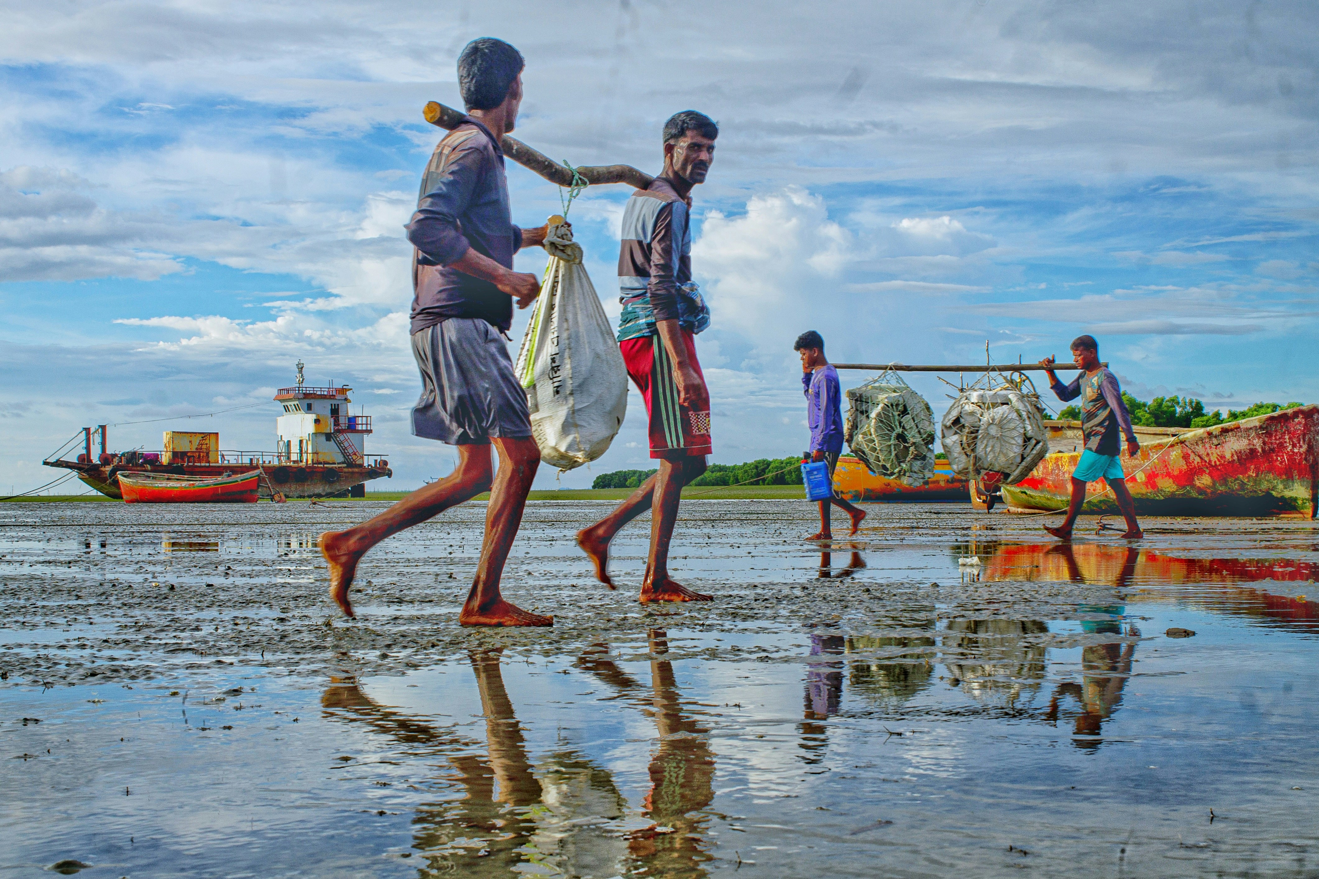 Four people carrying loads on poles across a muddy tidal flat near colorful boats