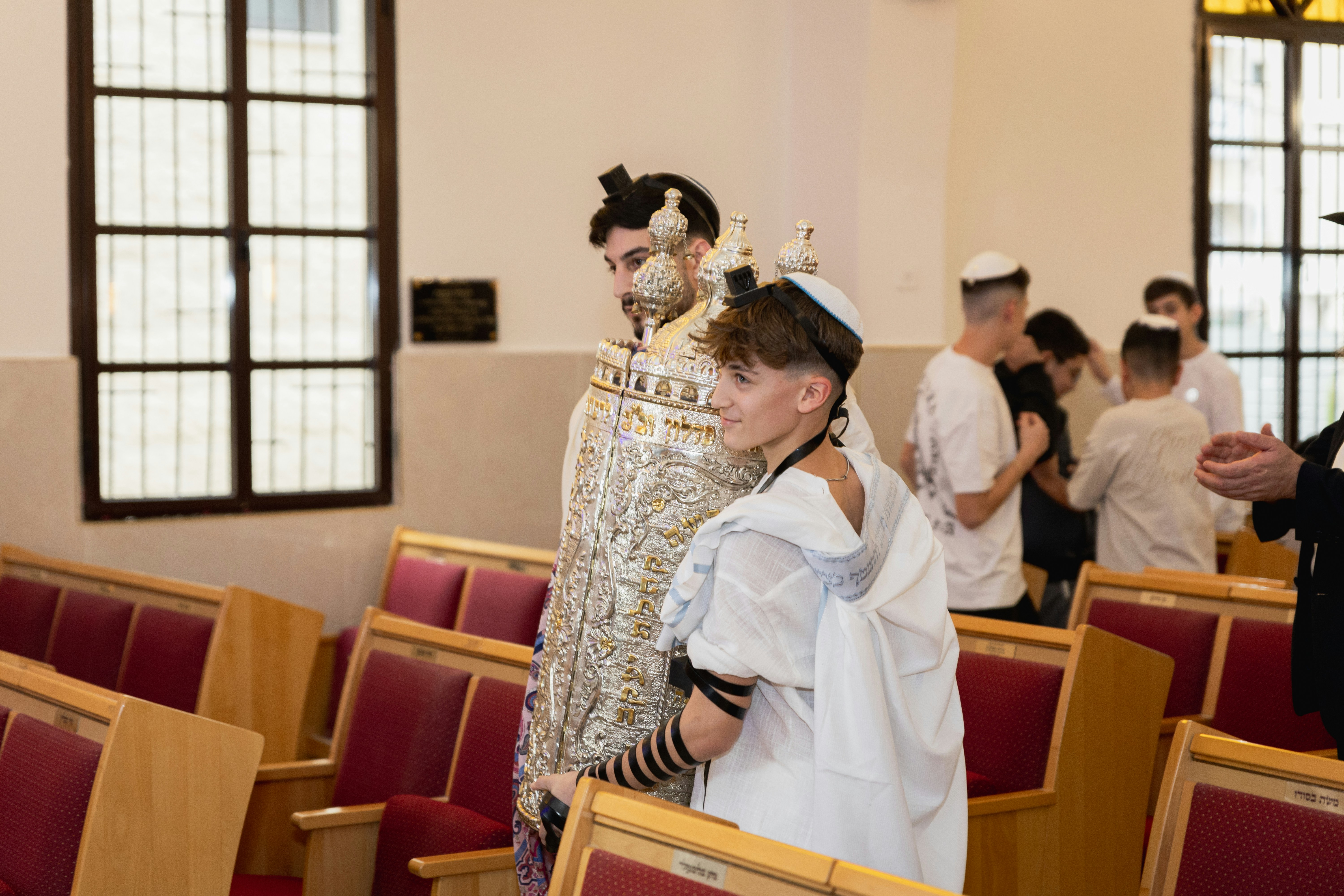 A young person wearing a tallit and tefillin carrying a silver Torah scroll in a synagogue