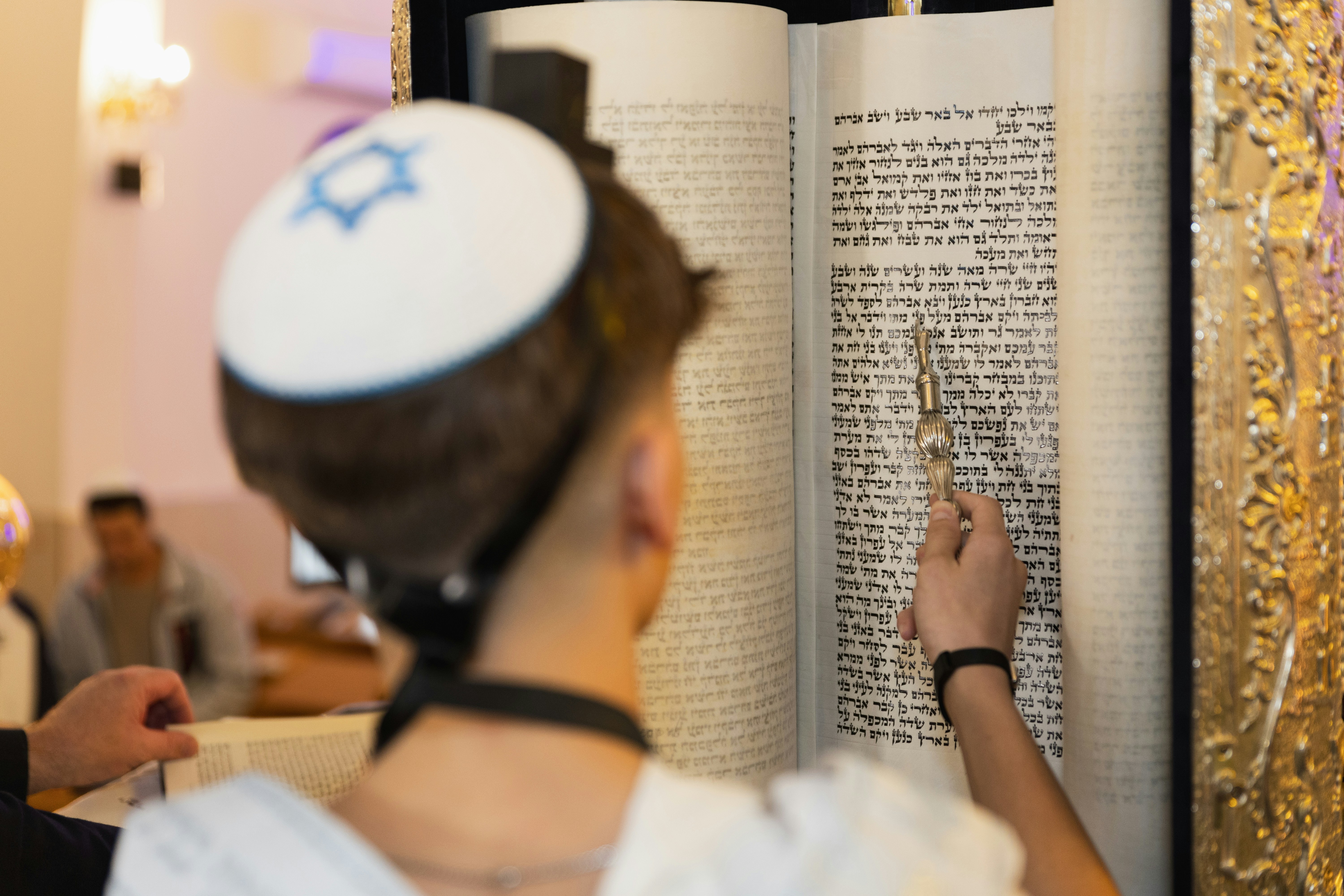 A person wearing a kippah reading from a Torah scroll with a silver pointer