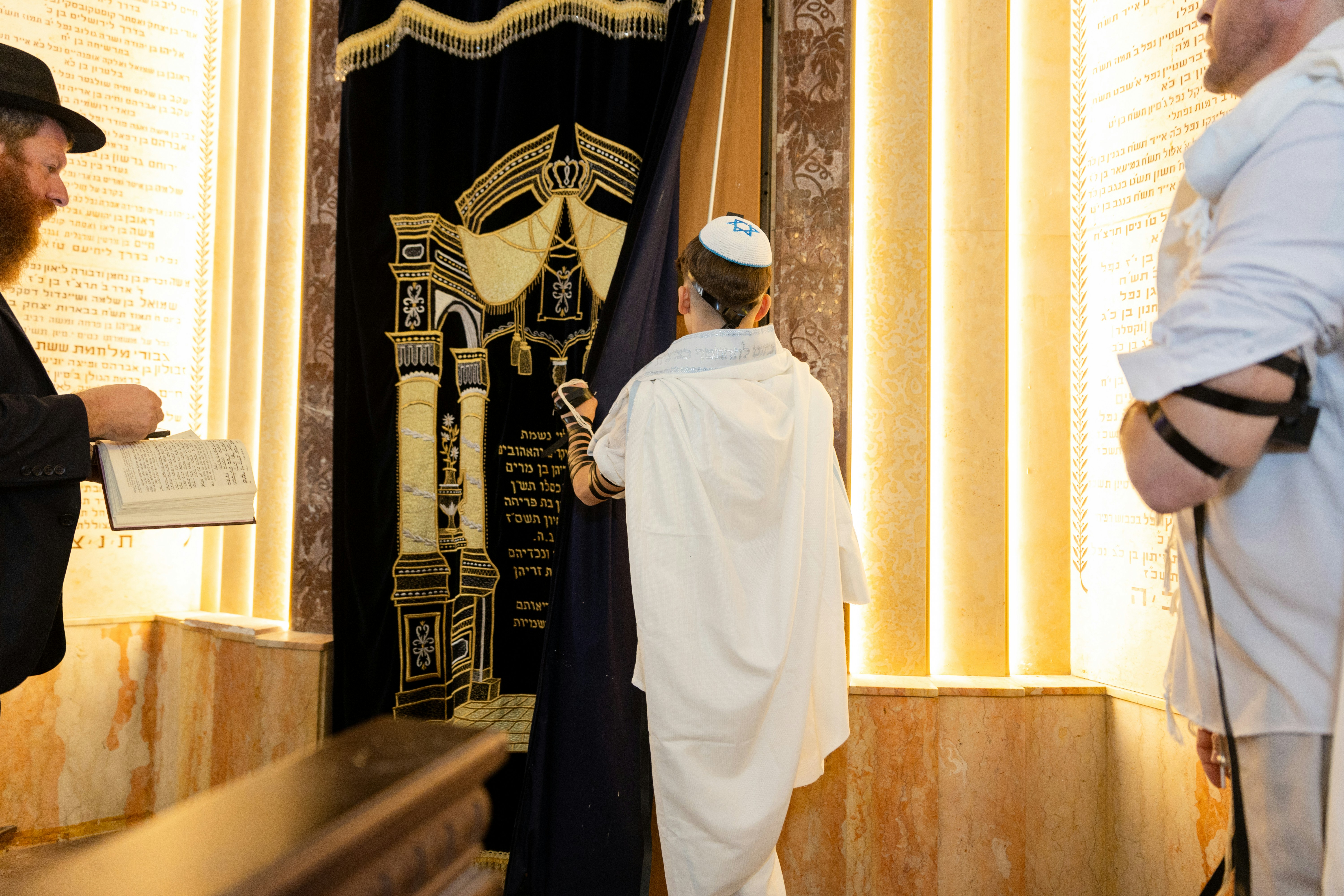 A boy in a tallit and kippah opening a dark curtain in a synagogue