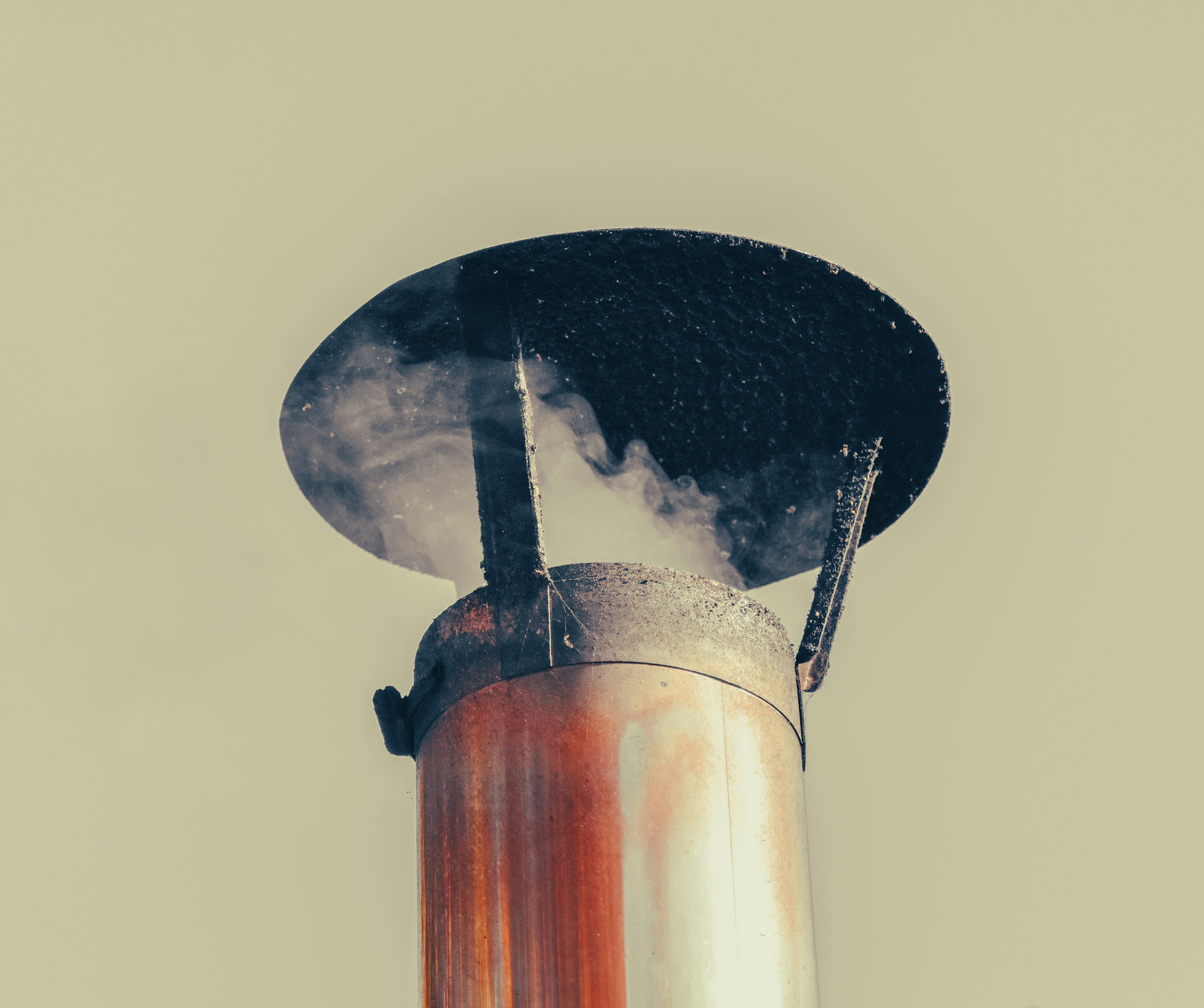Smoke rising from a metal chimney pipe with a circular cap against a pale sky