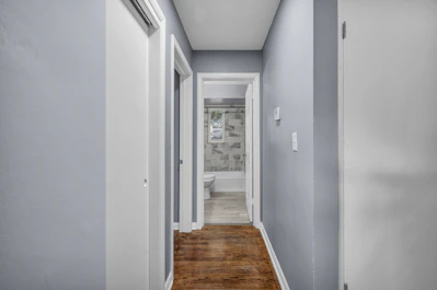 A narrow hallway with light blue walls and wooden flooring leading to a bathroom