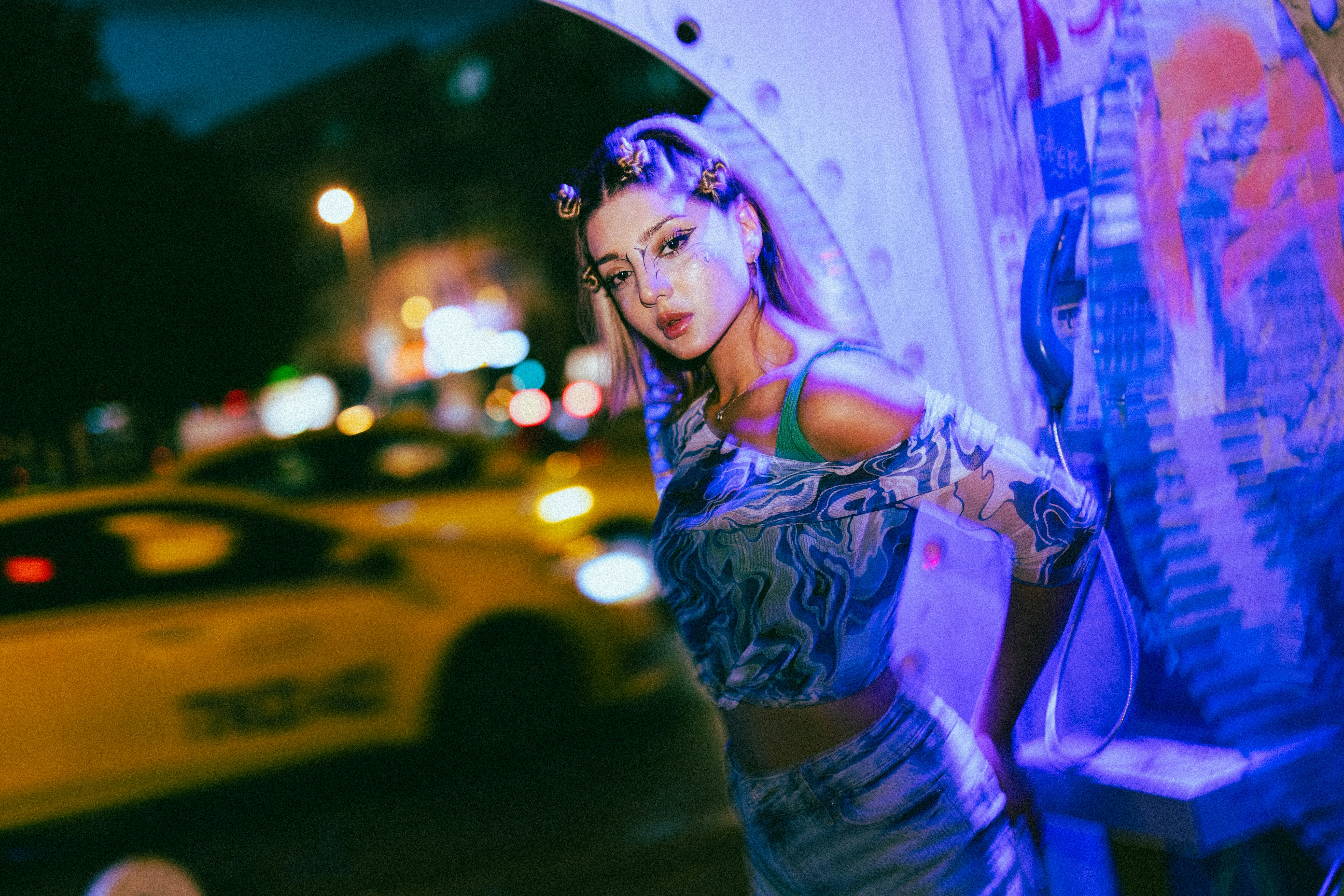 A woman in a patterned top standing by a phone booth on a city street at night