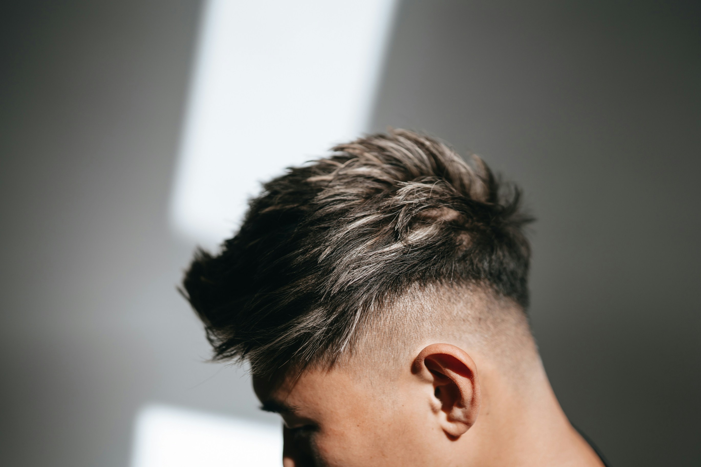 A person with a textured fade haircut in profile against a grey wall with light