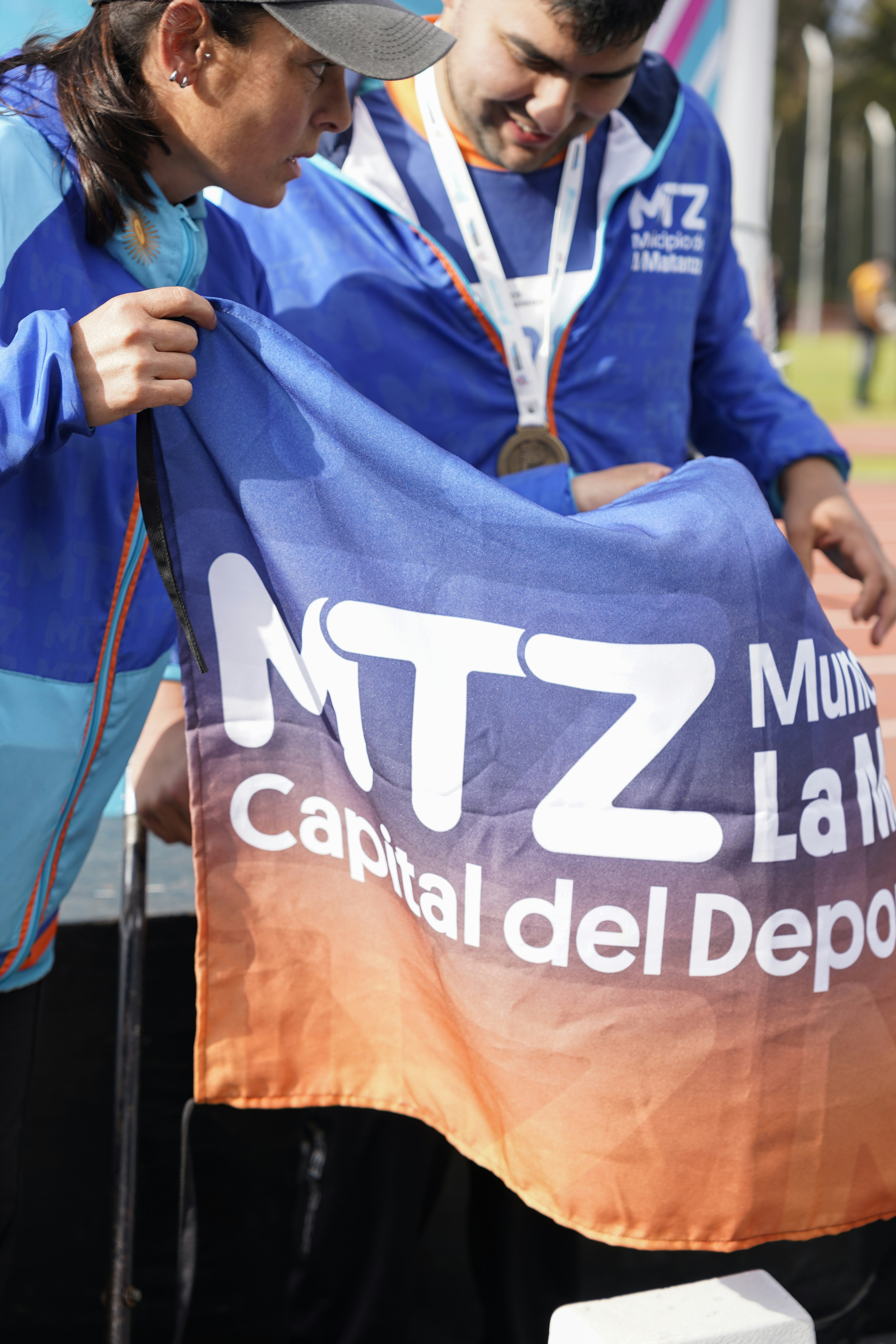 Two people in blue athletic jackets holding a flag with the text MTZ Capital del Deporte