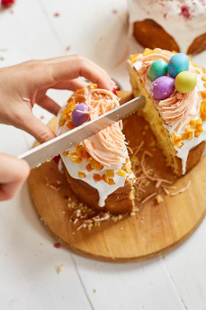 A person slicing a decorated Easter cake topped with icing, candied fruit, and colorful chocolate eggs