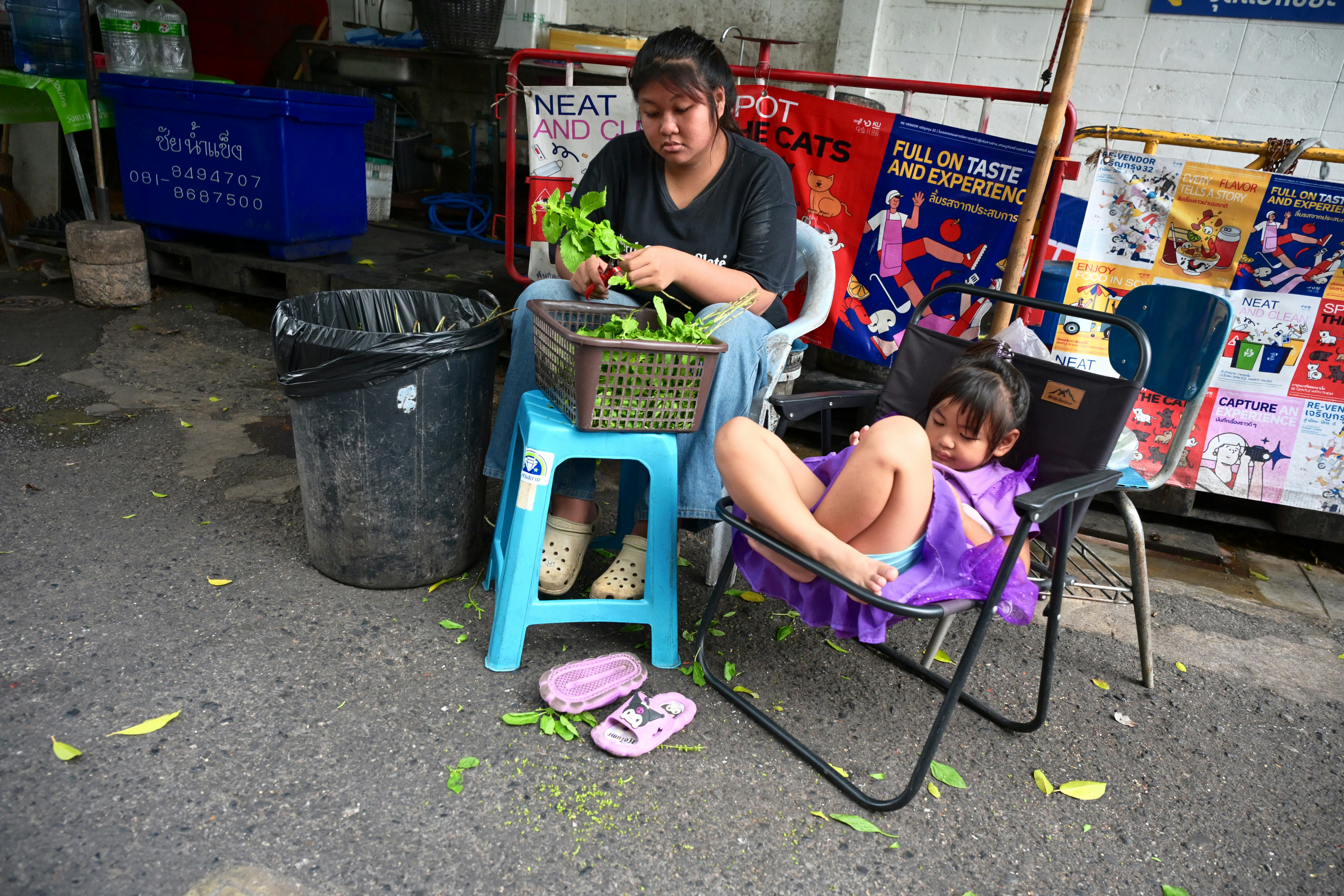 A woman trimming green herbs into a basket while a child rests in a folding chair