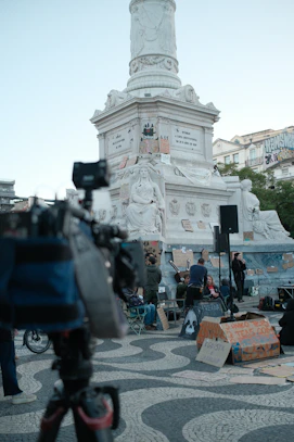 A camera on a tripod overlooking a monument with protest signs and people in Lisbon