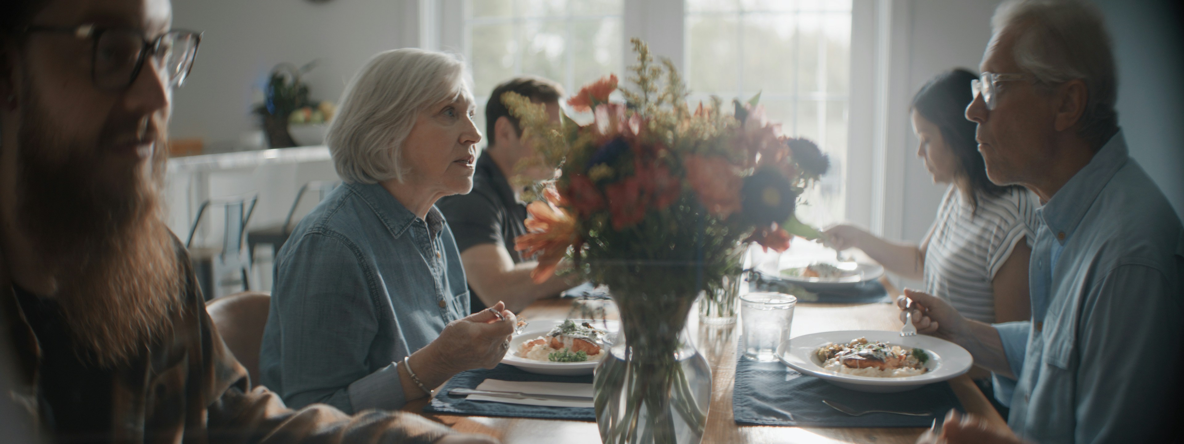 A family eating dinner together at a long wooden table with a floral centerpiece