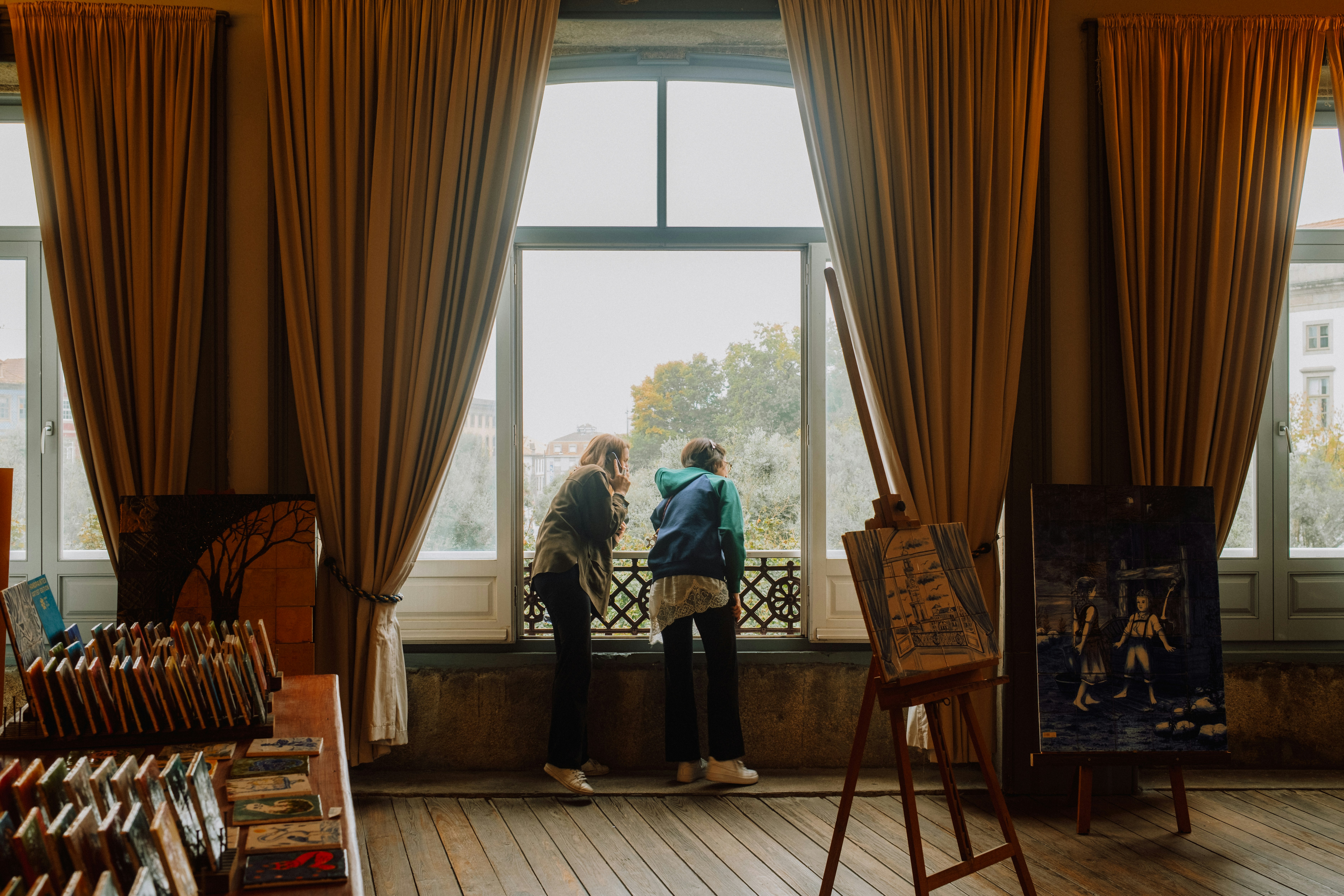 Two people looking out a large window in a room with art supplies and curtains