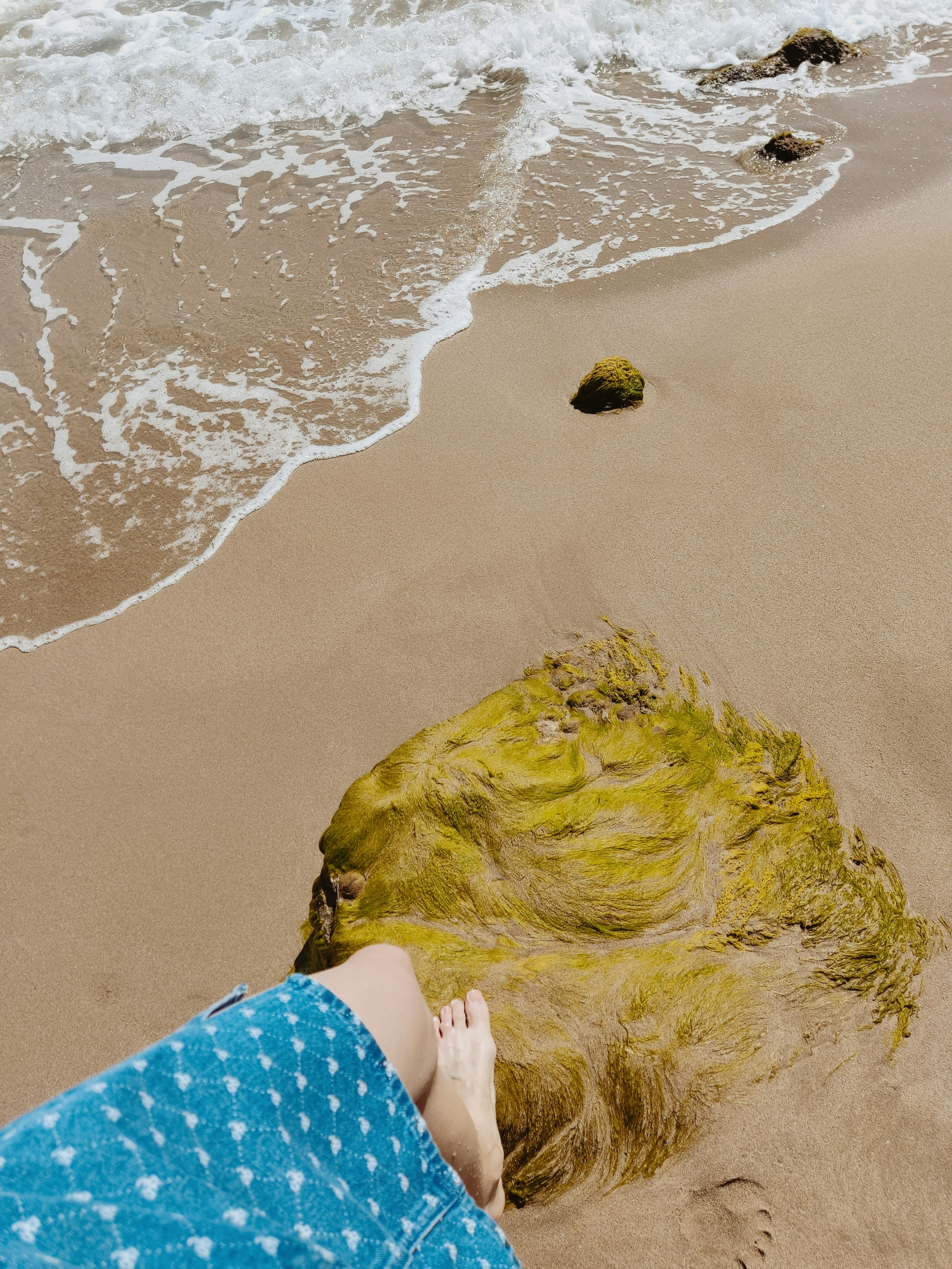 A person in a blue patterned skirt sitting on a mossy rock on a sandy beach