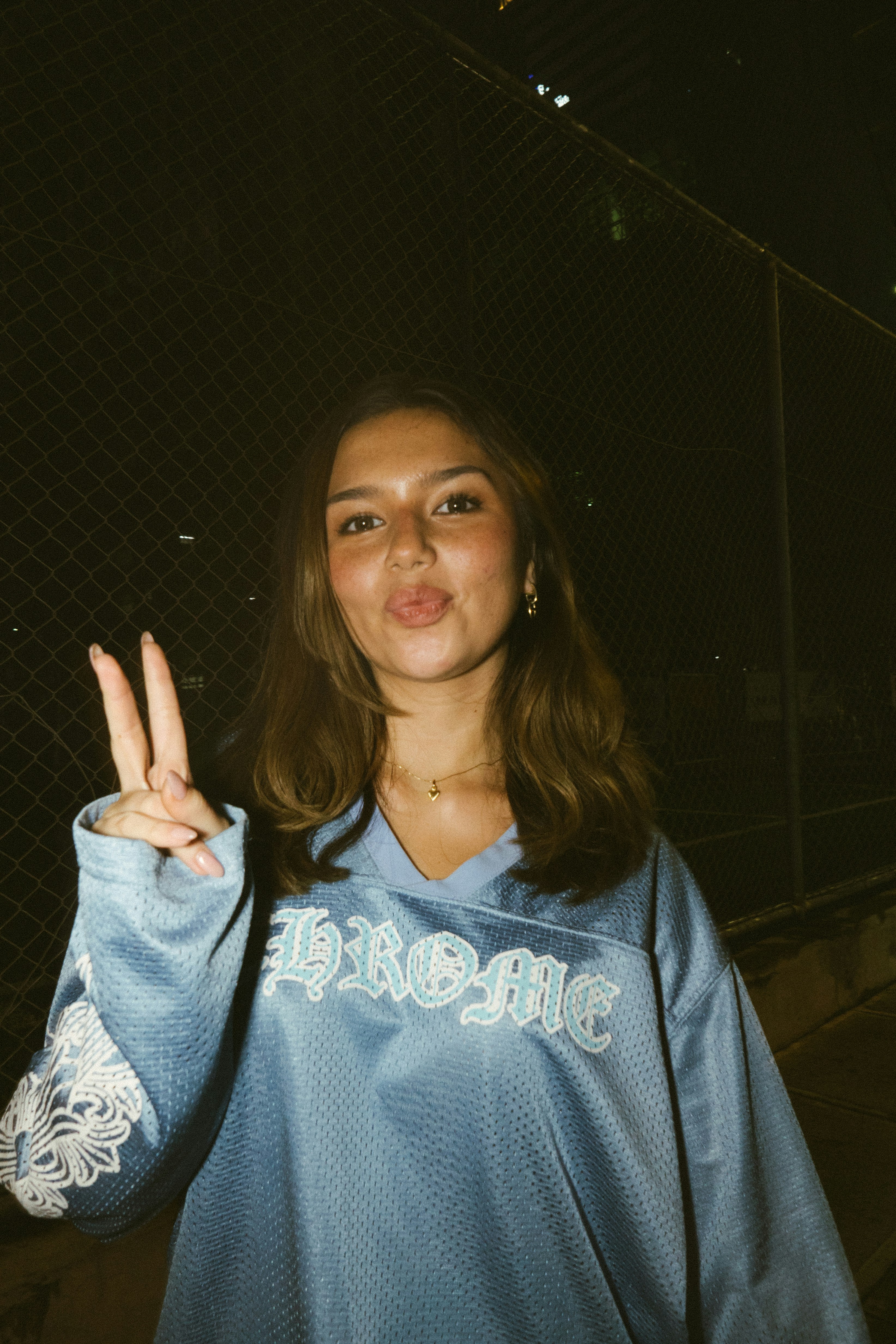 A young woman in a blue jersey making a peace sign against a chain-link fence at night