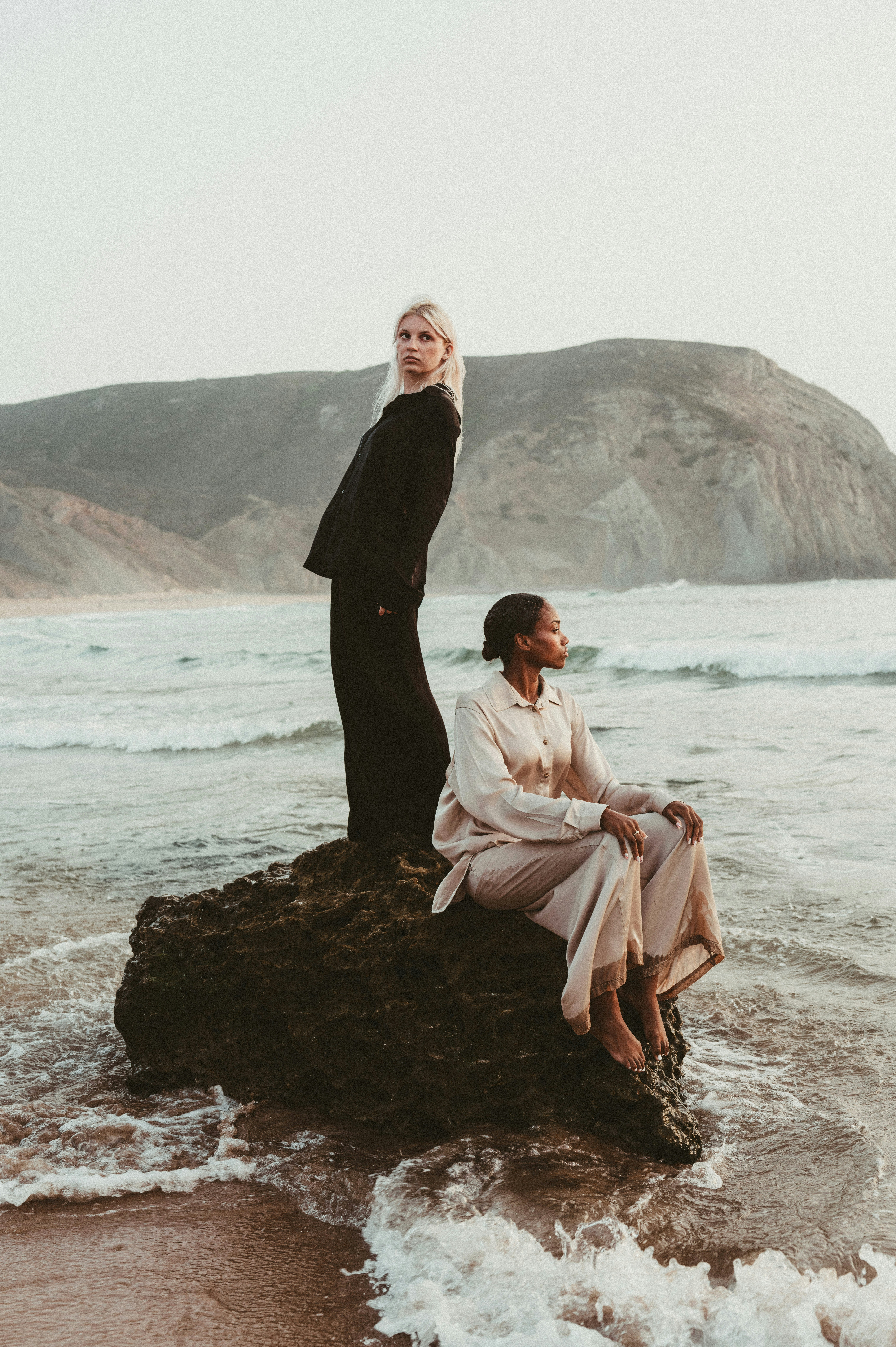 Two adults on a rock in the ocean, one standing and one sitting, at low sun - 相关推荐