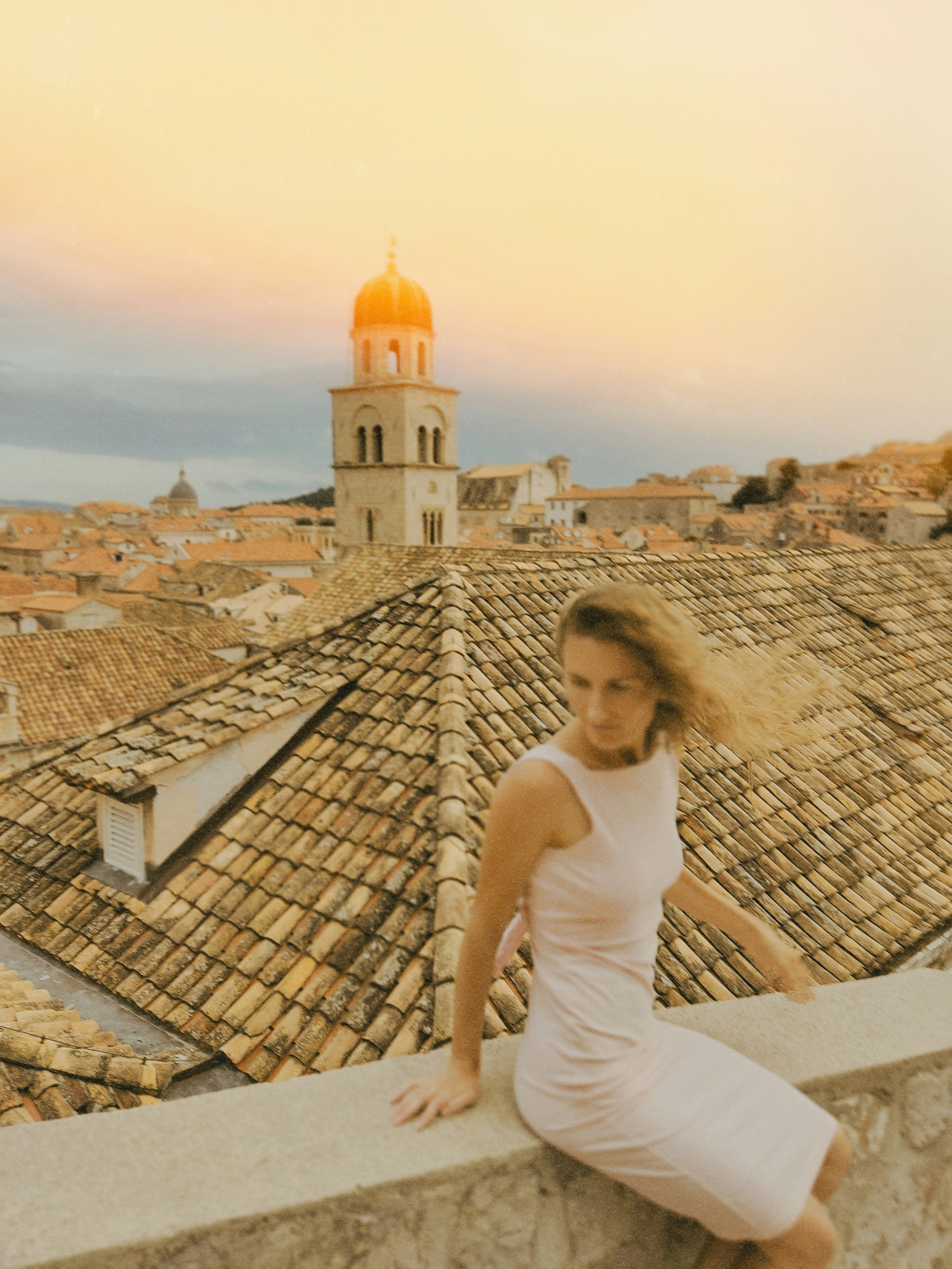 A woman in a light dress sitting on a stone wall overlooking Dubrovnik rooftops - 相关推荐