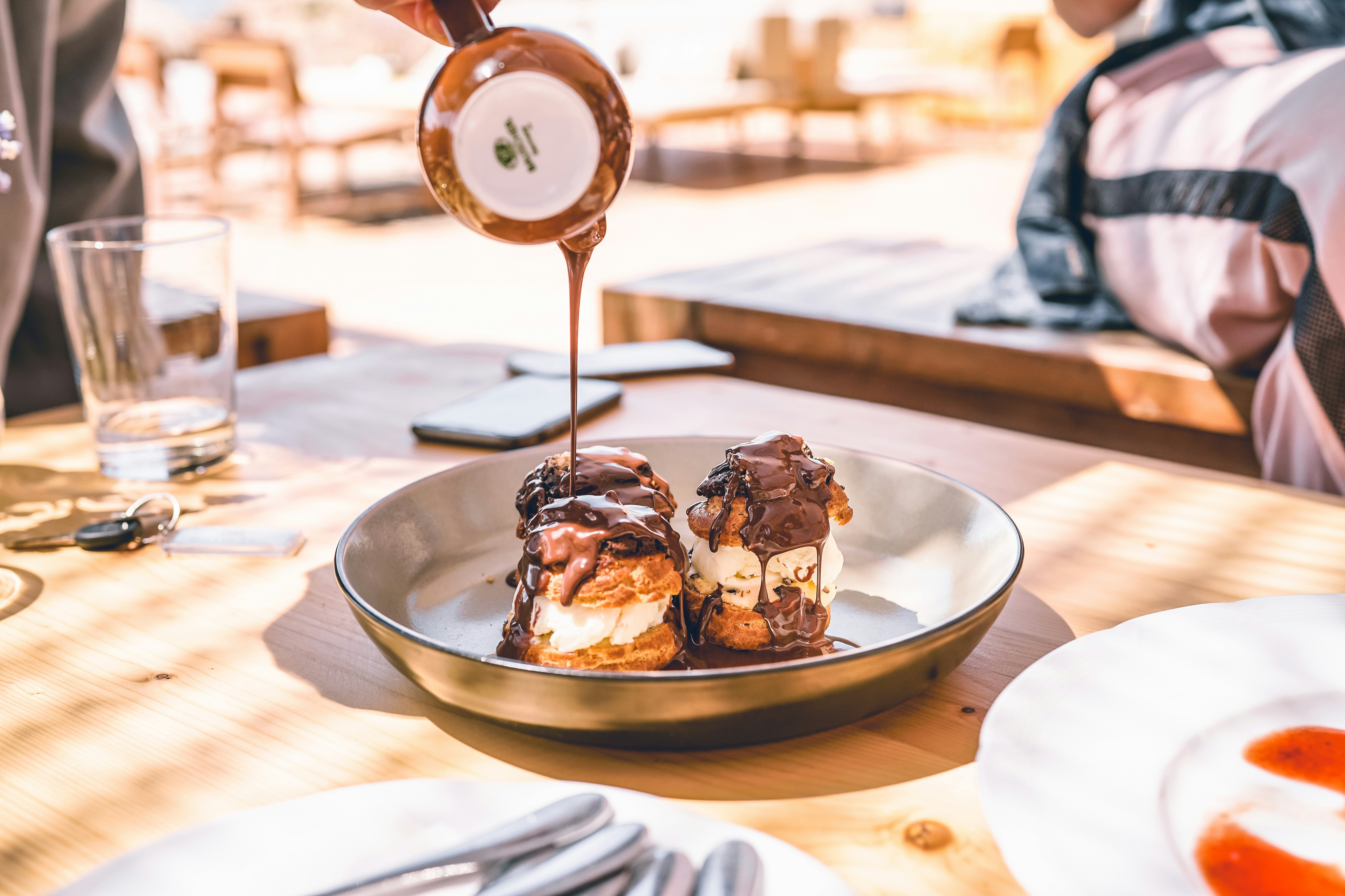 Chocolate sauce being poured from a small pitcher over two cream puffs on a plate