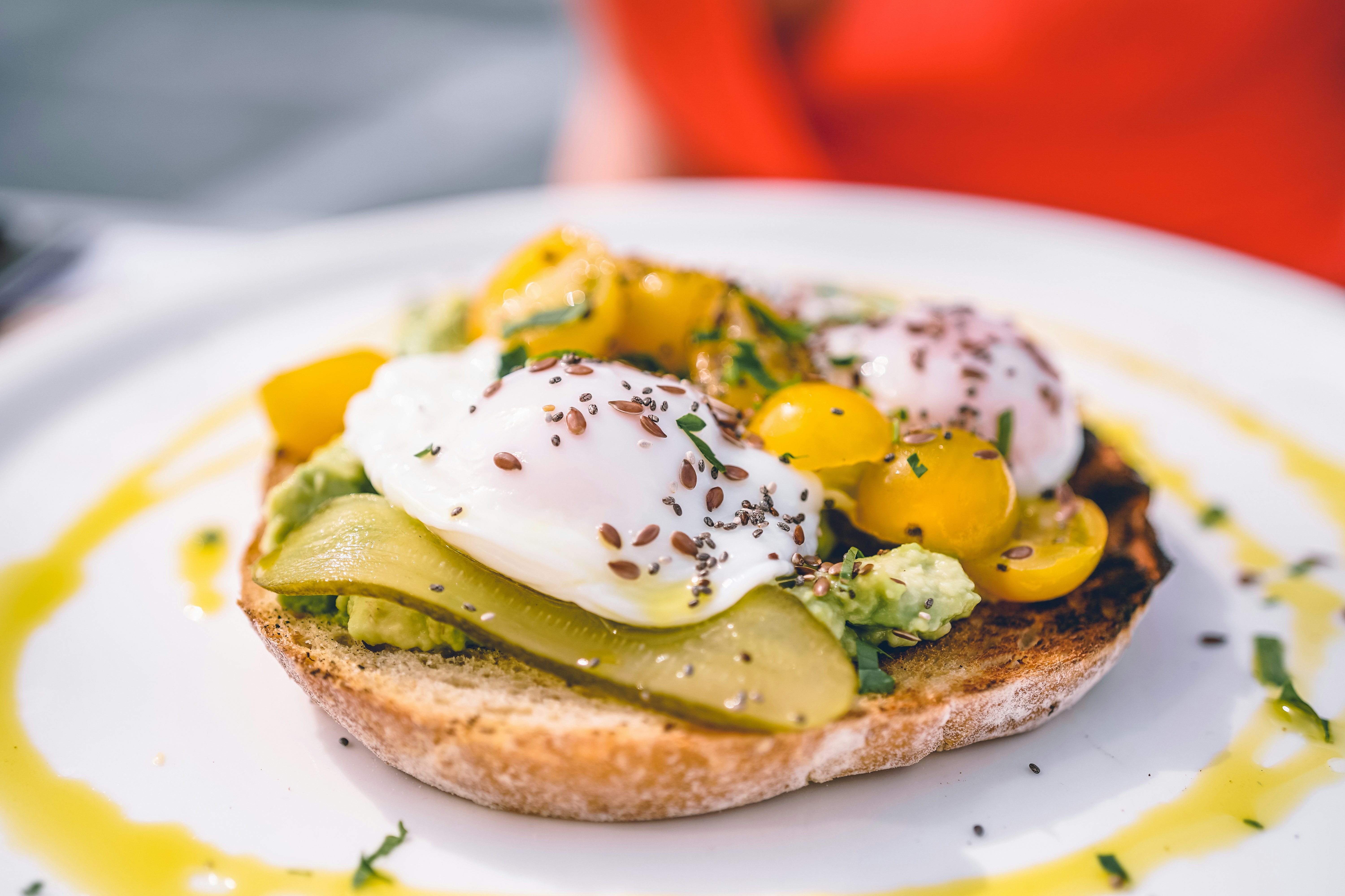 Poached eggs on avocado toast with cherry tomatoes, herbs and a swirl of olive oil, served on a white plate in the sun.