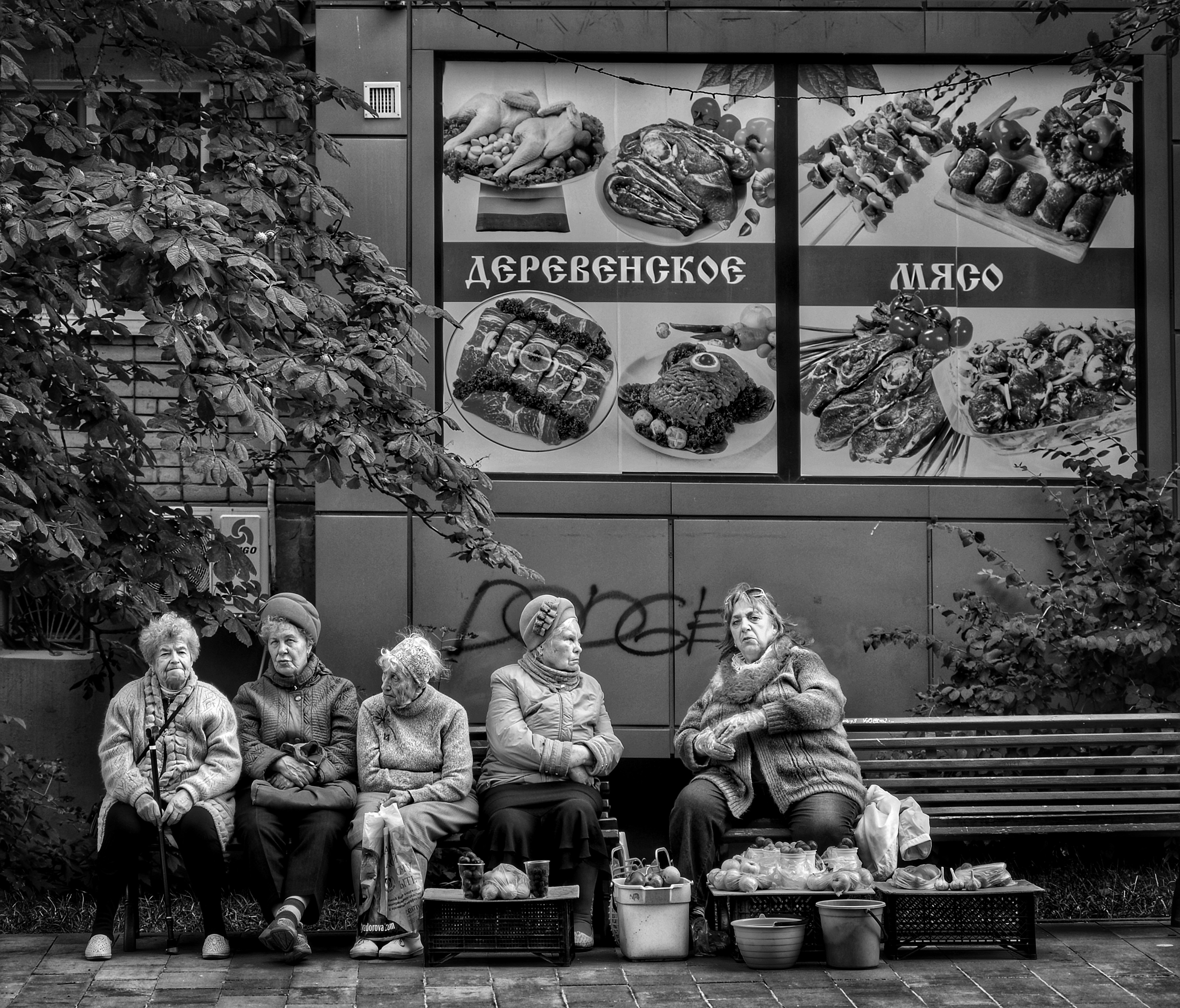 Five elderly women sitting on a bench in front of a meat shop advertisement
