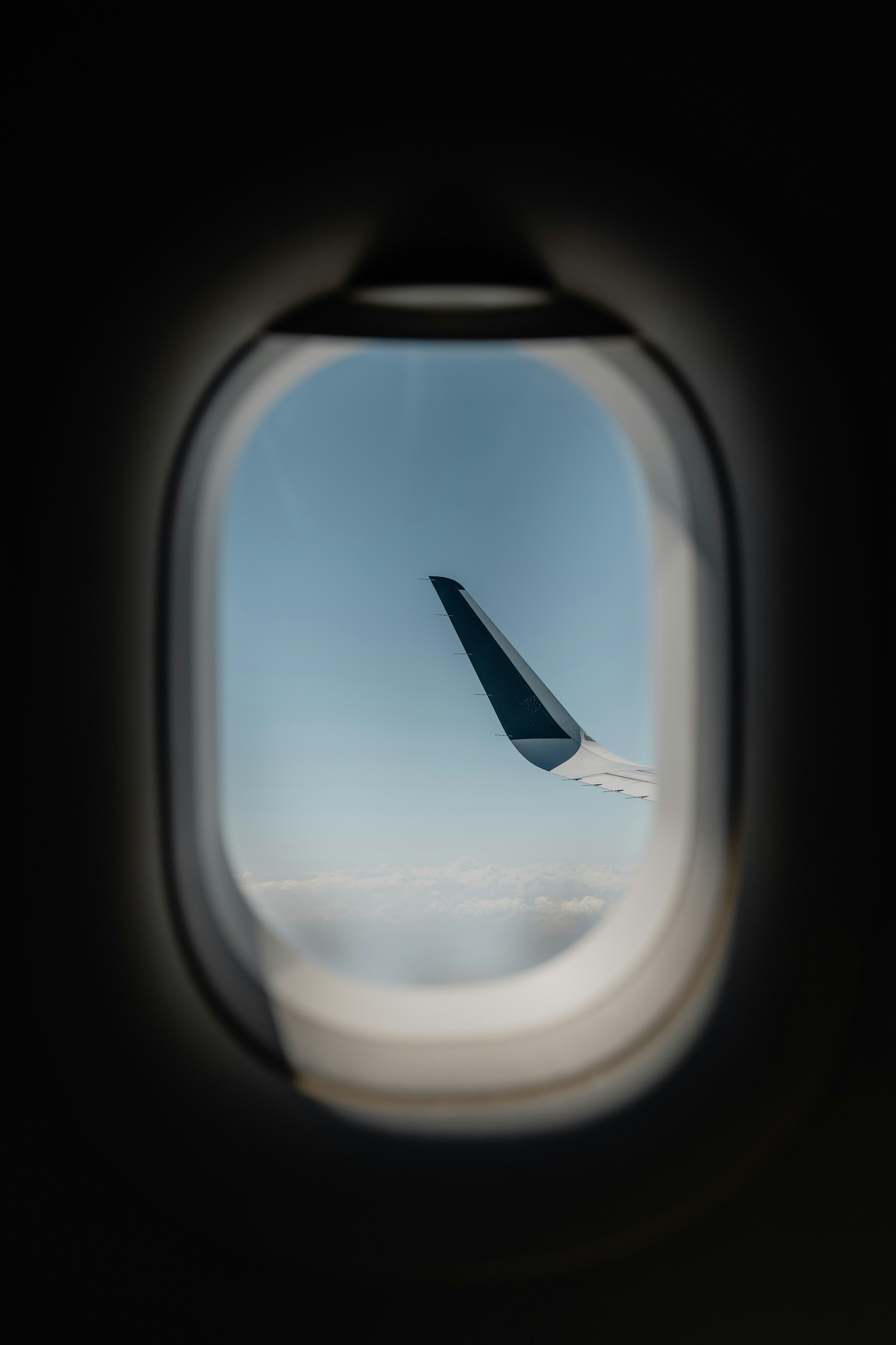 An airplane wing viewed through a dark oval cabin window against a clear blue sky