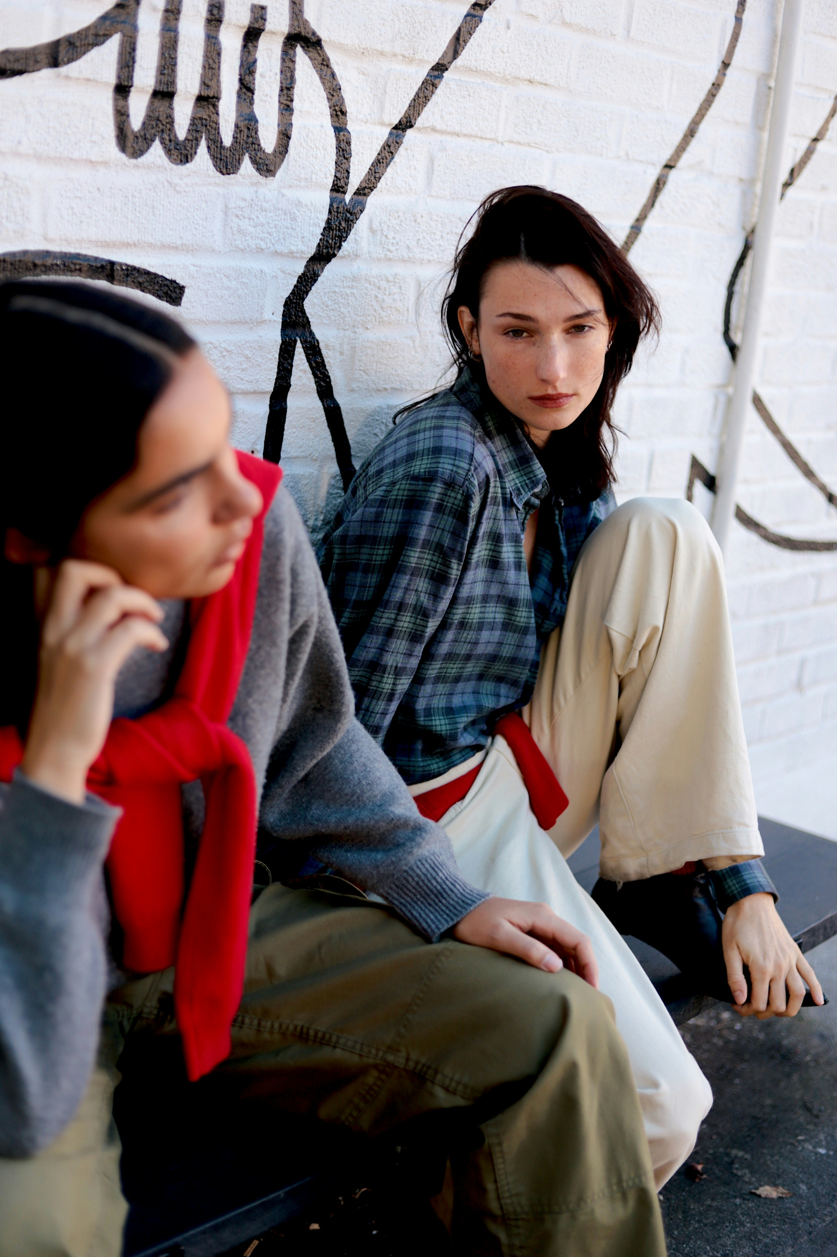 Two women sitting on a bench against a white brick wall with black graffiti
